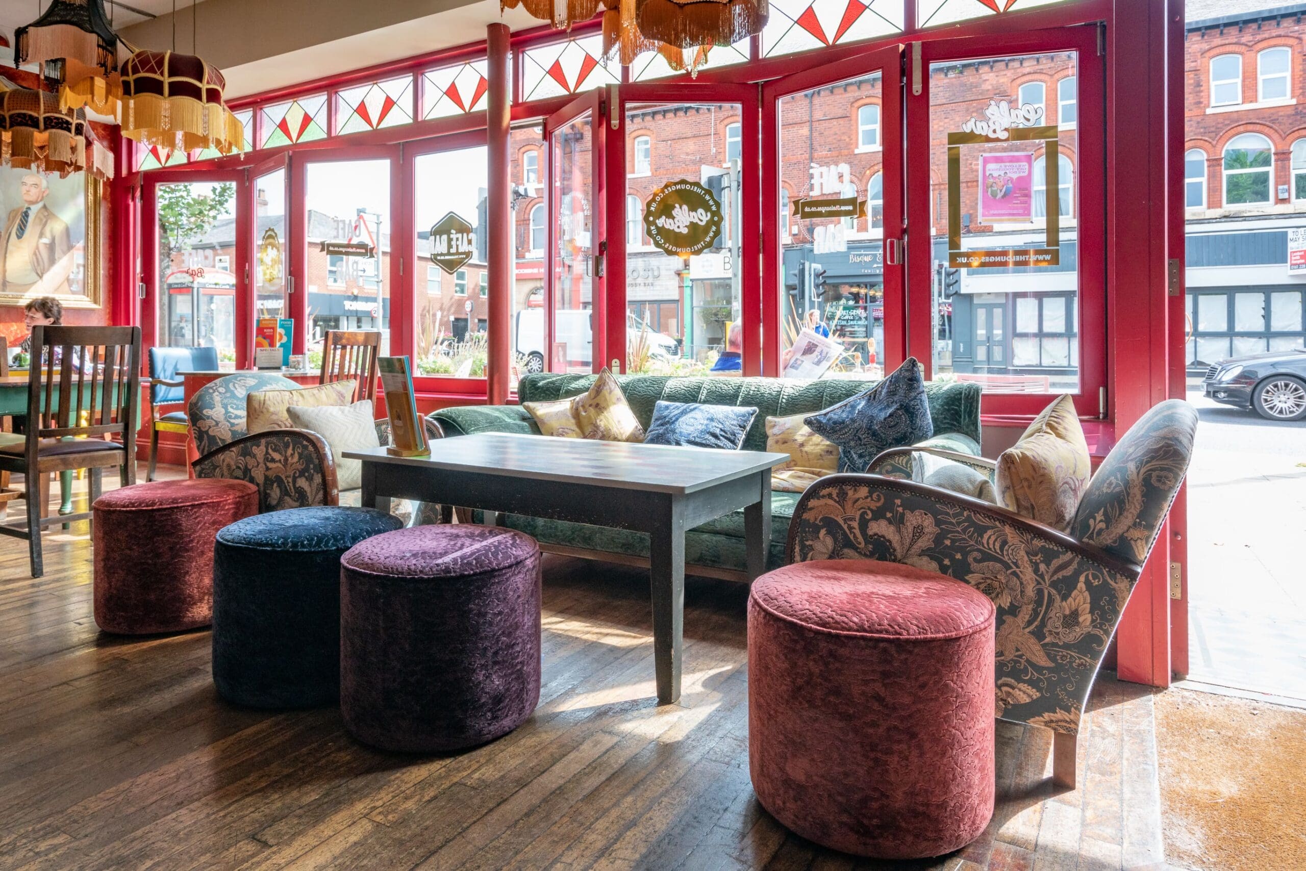 A cozy café interior, perfect for an Expo meetup, features colorful plush stools, patterned armchairs, a wooden table, and a green sofa set against large red-framed windows with daylight streaming in and a lively street view outside.