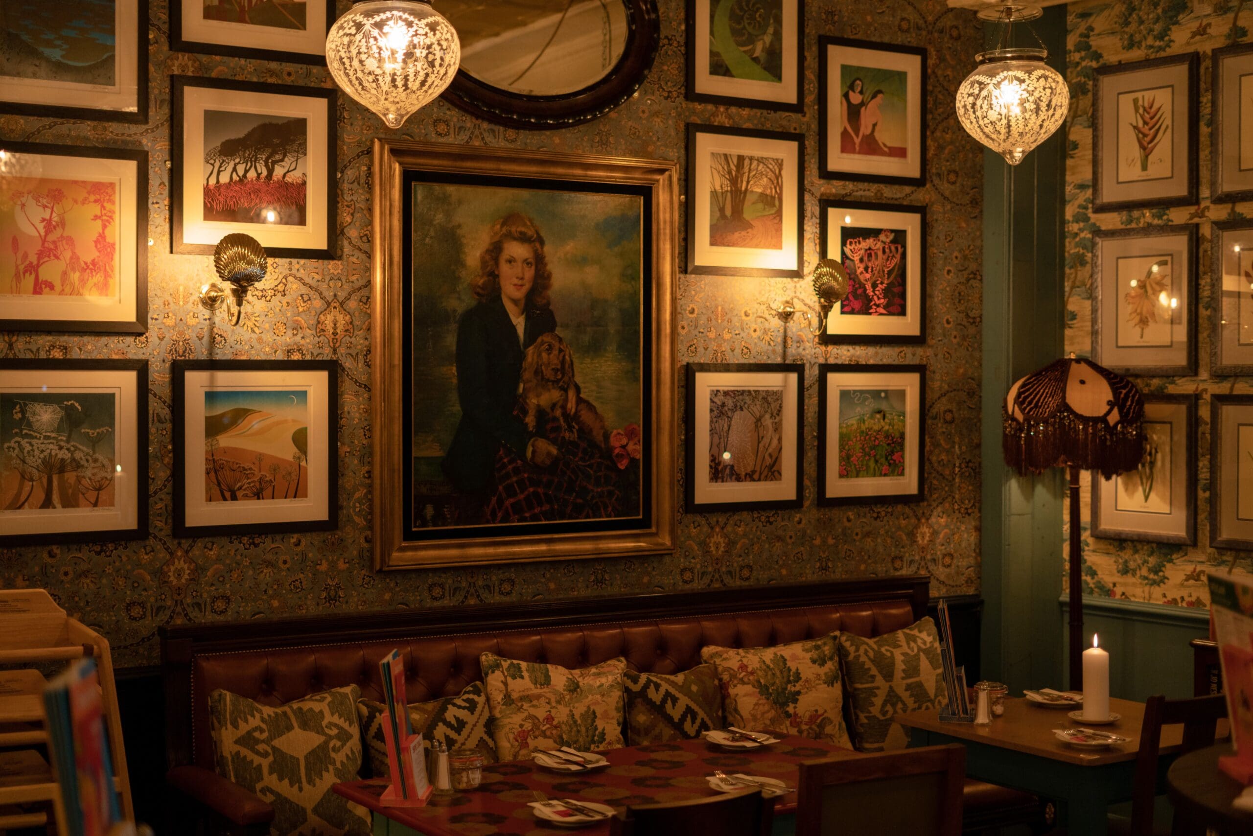 A cozy, dimly lit restaurant with patterned wallpaper, eclectic artwork and framed pictures on the walls, a vintage portrait above plush pillows on a leather bench, and ornate chandeliers casting a warm glow near the quaint fosso.