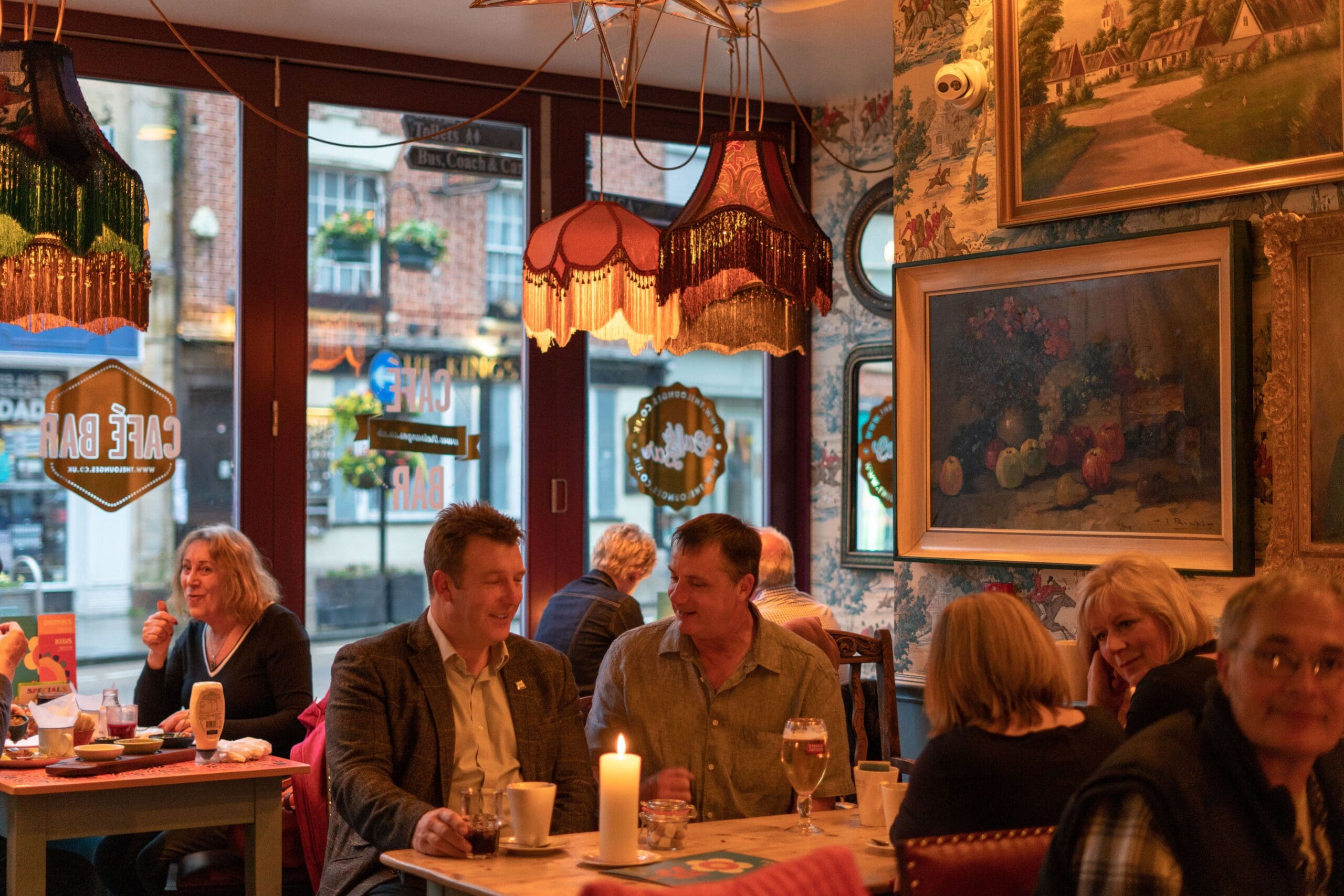 A cozy café with vintage décor, warm lighting, and several people sitting at tables by the window, enjoying drinks and conversation. Framed paintings and patterned wallpaper decorate the walls, giving the fosso a welcoming atmosphere.