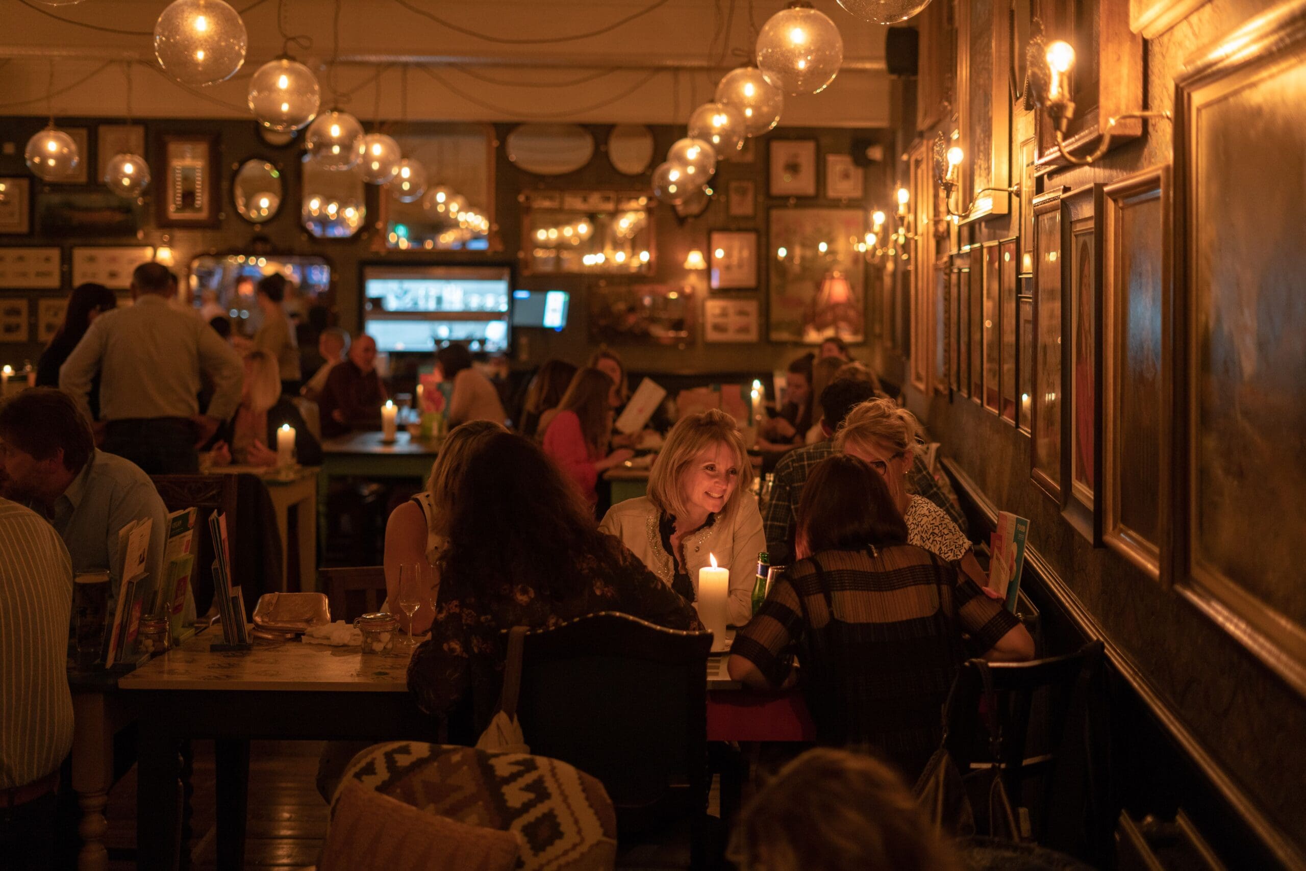A warmly lit restaurant filled with people dining, chatting, and enjoying themselves. Tables are decorated with candles, while fosso-inspired framed pictures and glowing lights on the walls create a cozy, inviting atmosphere.