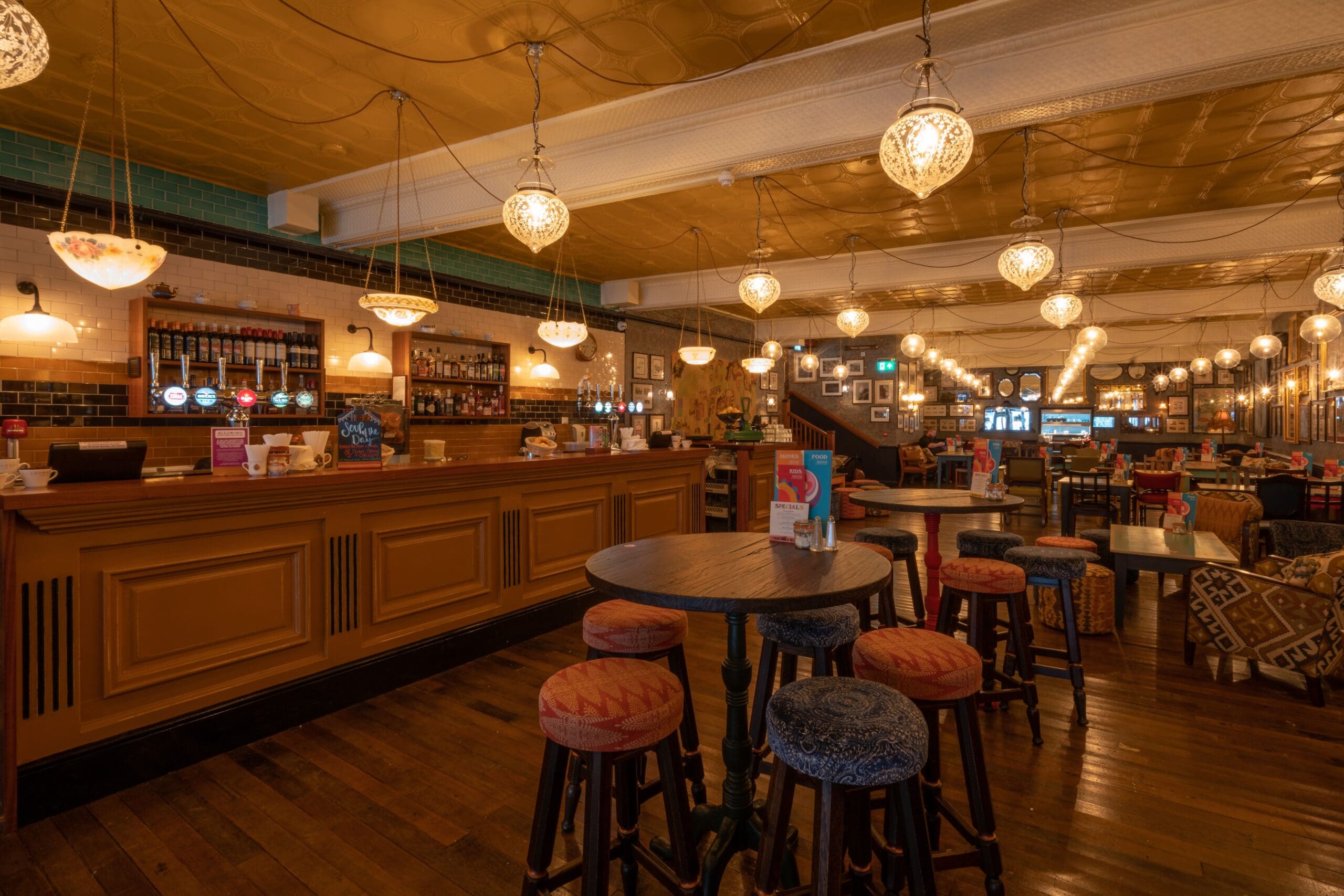 A warmly lit pub interior with round tables and cushioned stools, a long wooden bar beside a cozy fosso, hanging globe lights, framed pictures on the walls, and a polished wooden floor.