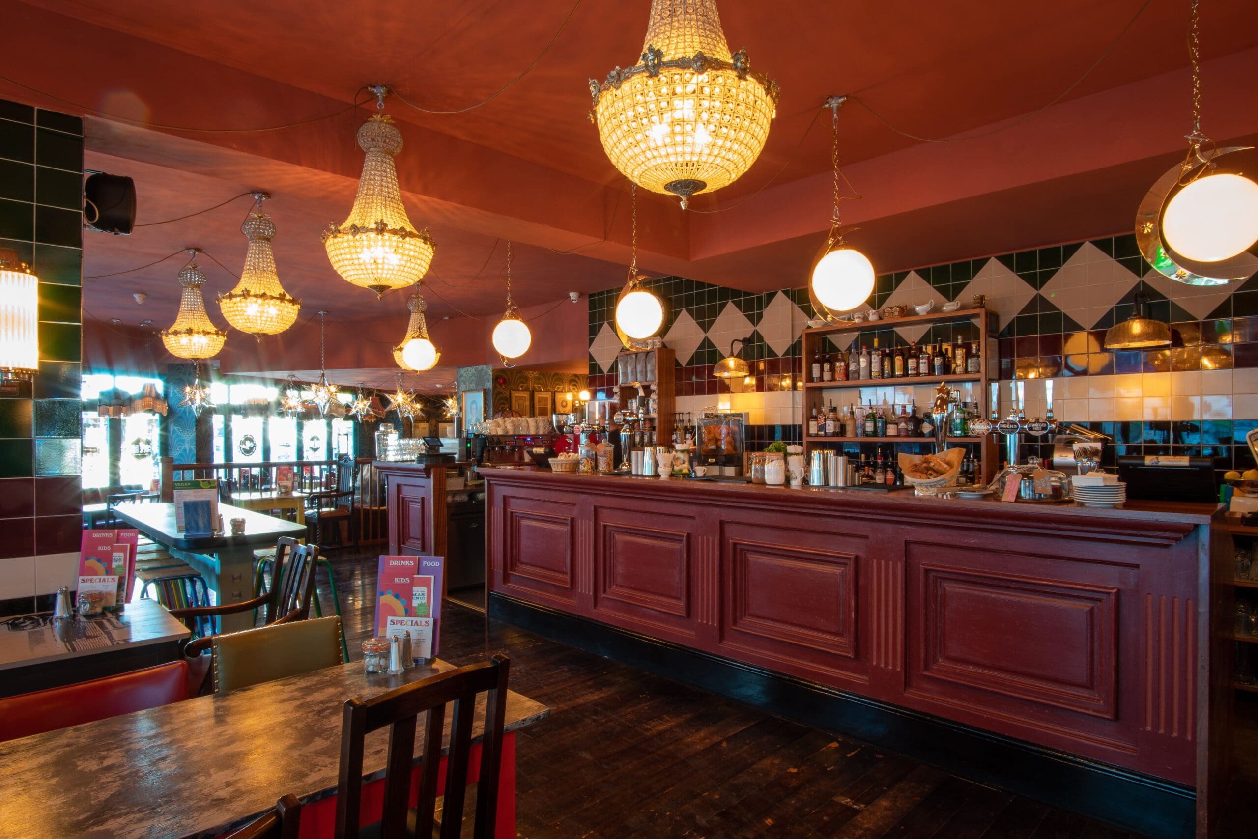 A cozy Marciano restaurant interior featuring ornate chandeliers, round globe lights, a long wooden bar with drinks and glasses, shelves filled with bottles, and tables with chairs on a polished dark wood floor.