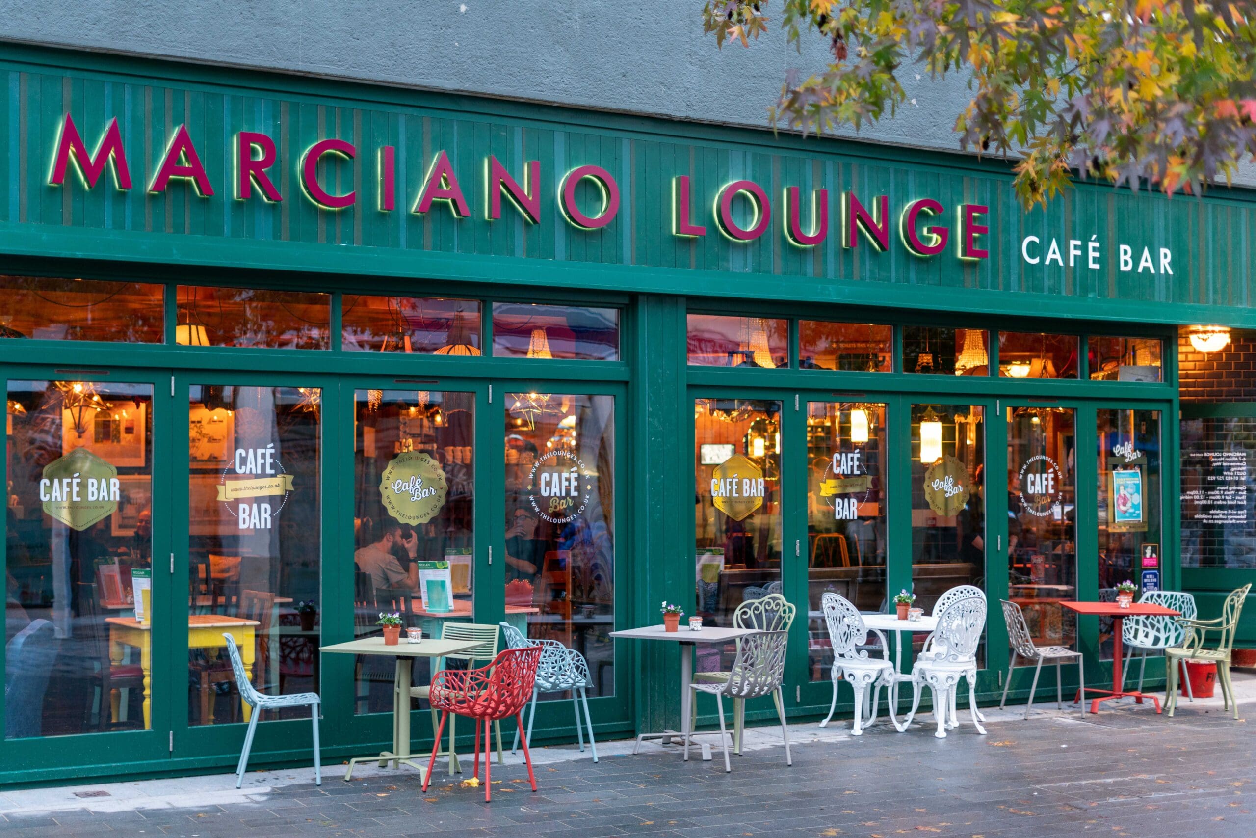 Marciano Lounge Café Bar exterior with green-framed windows and bold Marciano red signage. Outdoor seating features colorful chairs and tables, while warm indoor lighting and tree branches overhead create a welcoming atmosphere.