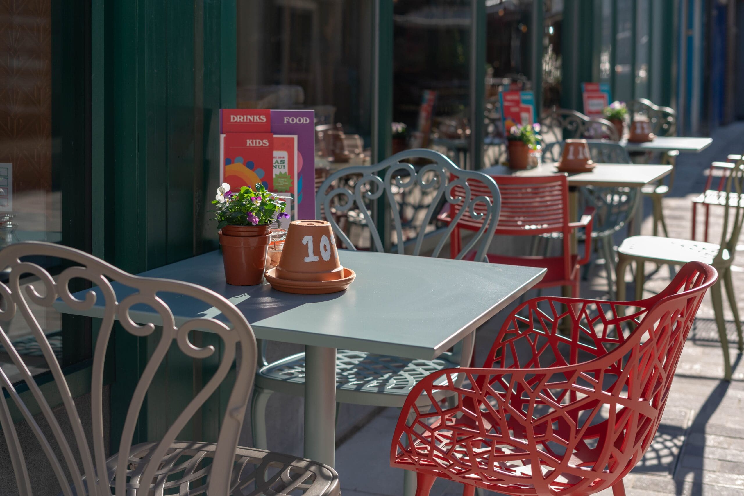 Outdoor café tables and chairs by Marciano, each with small flowerpots and numbered terracotta pots. Sunlight casts shadows, and colorful menus are displayed on the tables near a green-framed window.