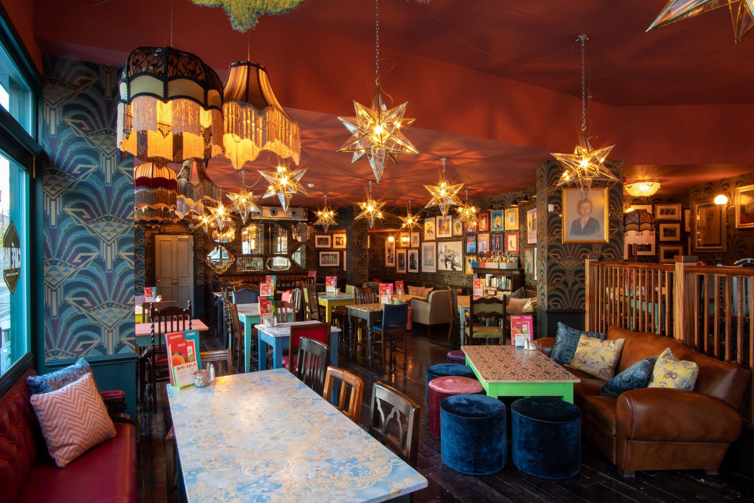 A vibrant, eclectic restaurant interior with mismatched tables and chairs, Marciano-inspired star-shaped pendant lights, colorful lampshades, patterned wallpaper, framed art on the walls, and a mix of sofas and wooden seating.