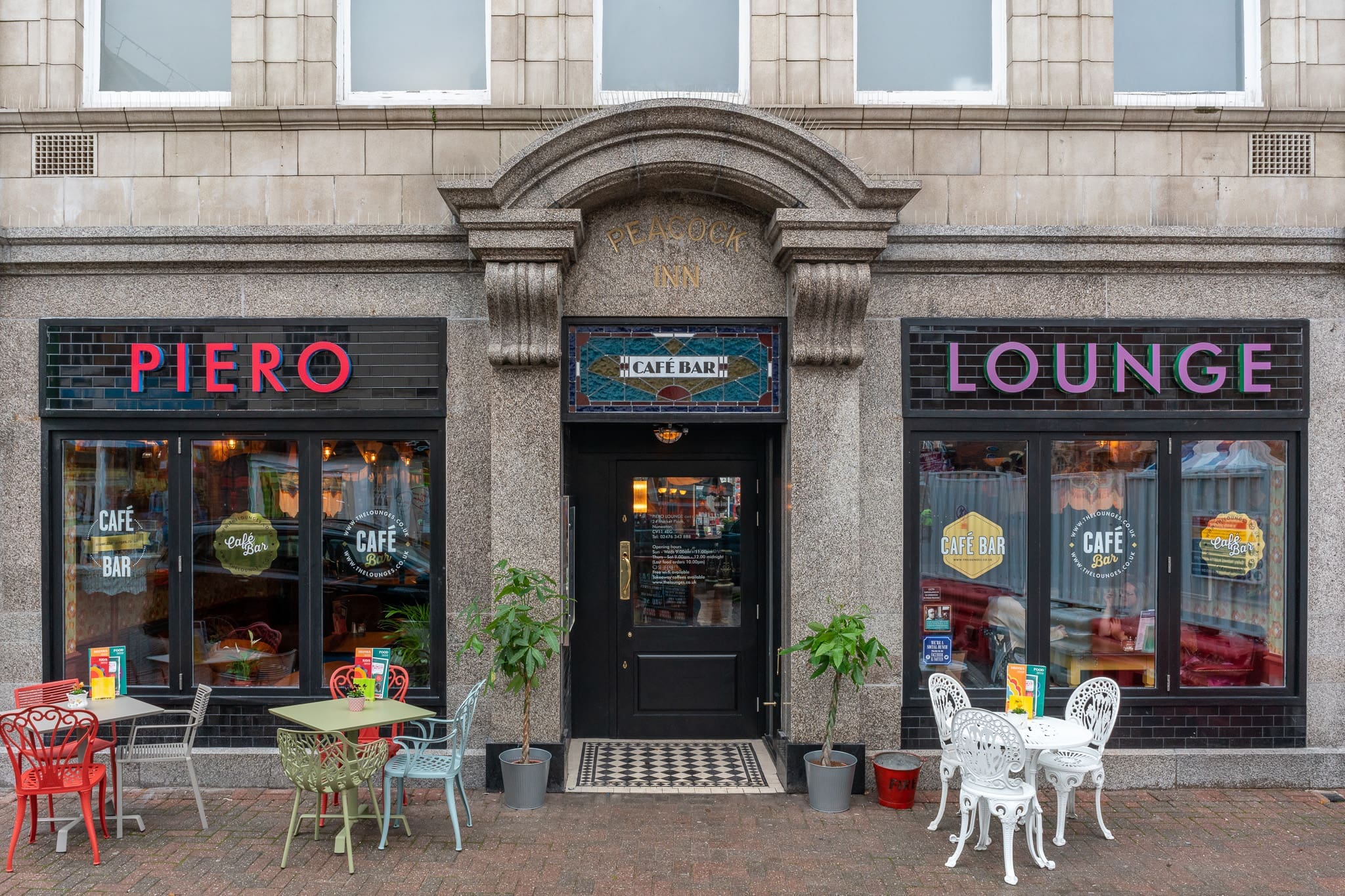 Street view of Piero café bar with two outdoor tables and chairs, bright signage reading "PIERO" and "LOUNGE," and large windows showing a colorful, cozy interior. The entrance is framed by an arch and decorative stonework.