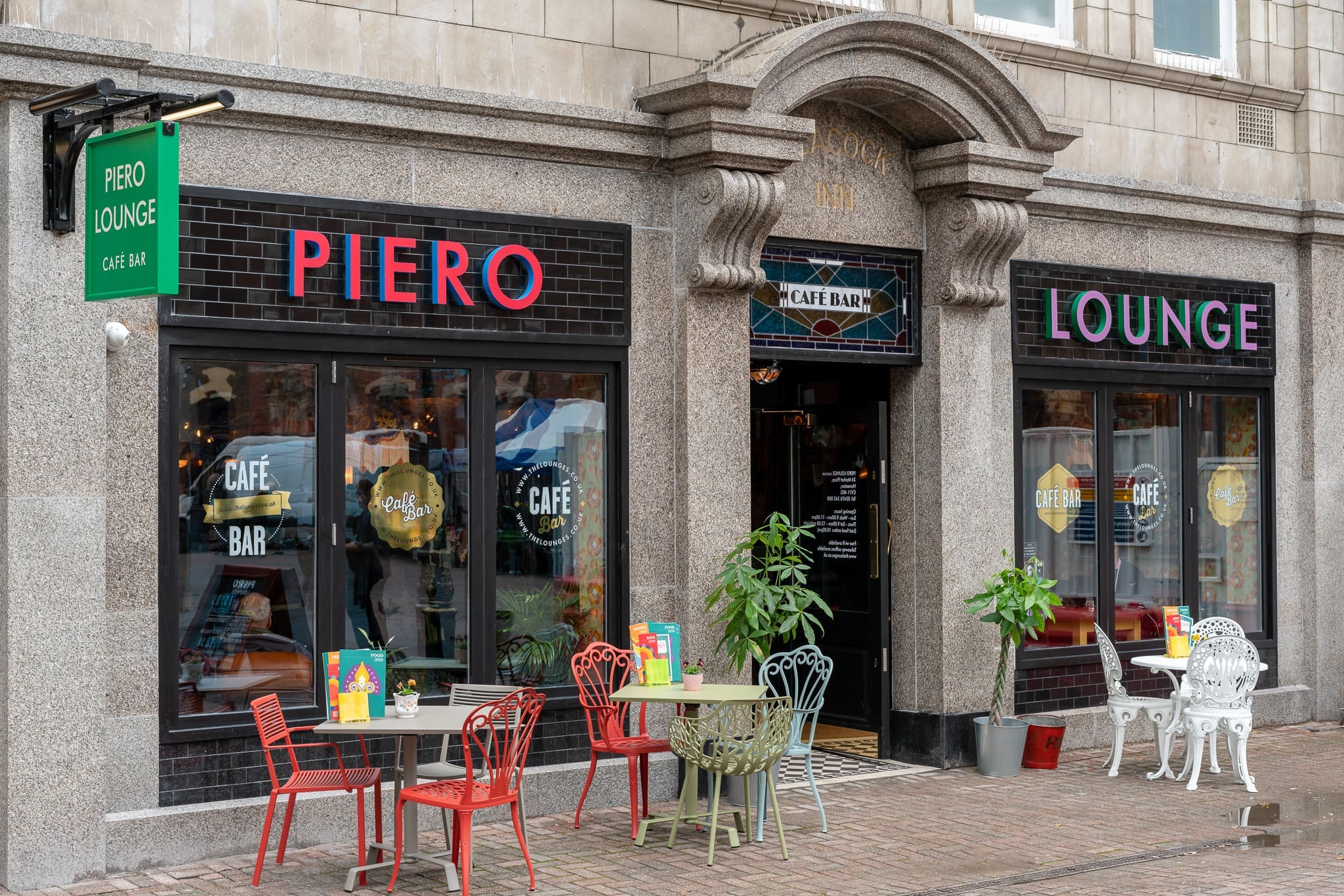 Piero Lounge café bar features a black-tiled exterior, large windows, colorful outdoor tables and chairs, potted plants, and playful menus on the tables—all set along a charming cobblestone street.