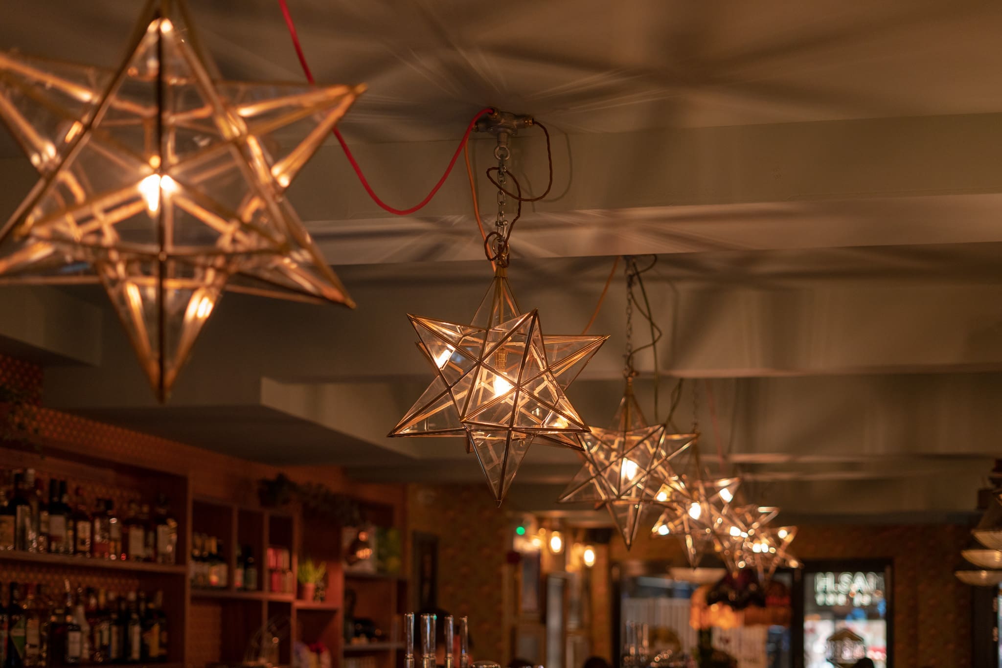 Hanging star-shaped pendant lights by Piero illuminate a cozy bar with shelves of bottles and glasses in the background, creating a warm and inviting atmosphere.