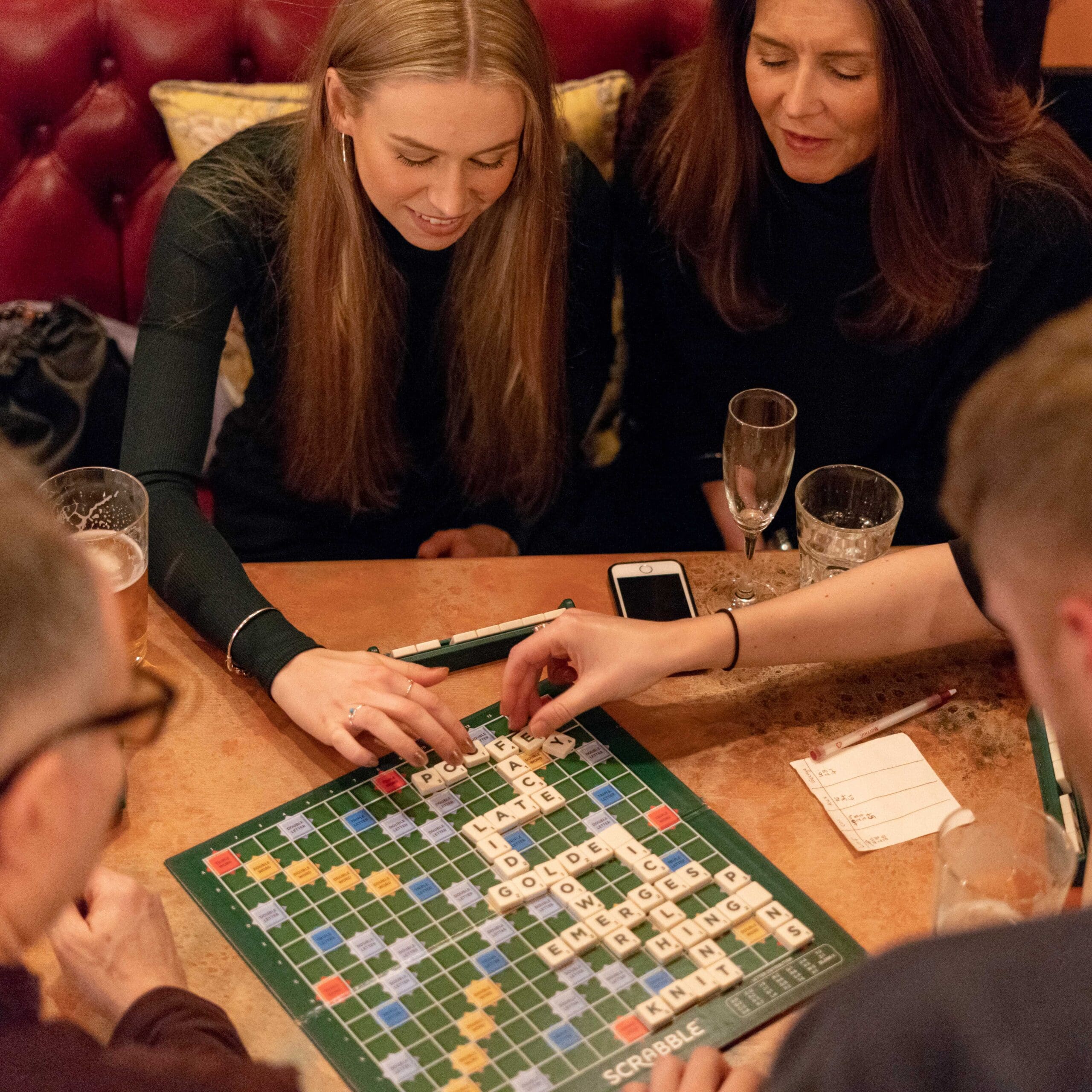 Four people sit around a table playing Scrabble, drinks—including Portello—nearby. Two women concentrate on the game while two others’ hands place tiles. A notepad and phone rest beside the board.