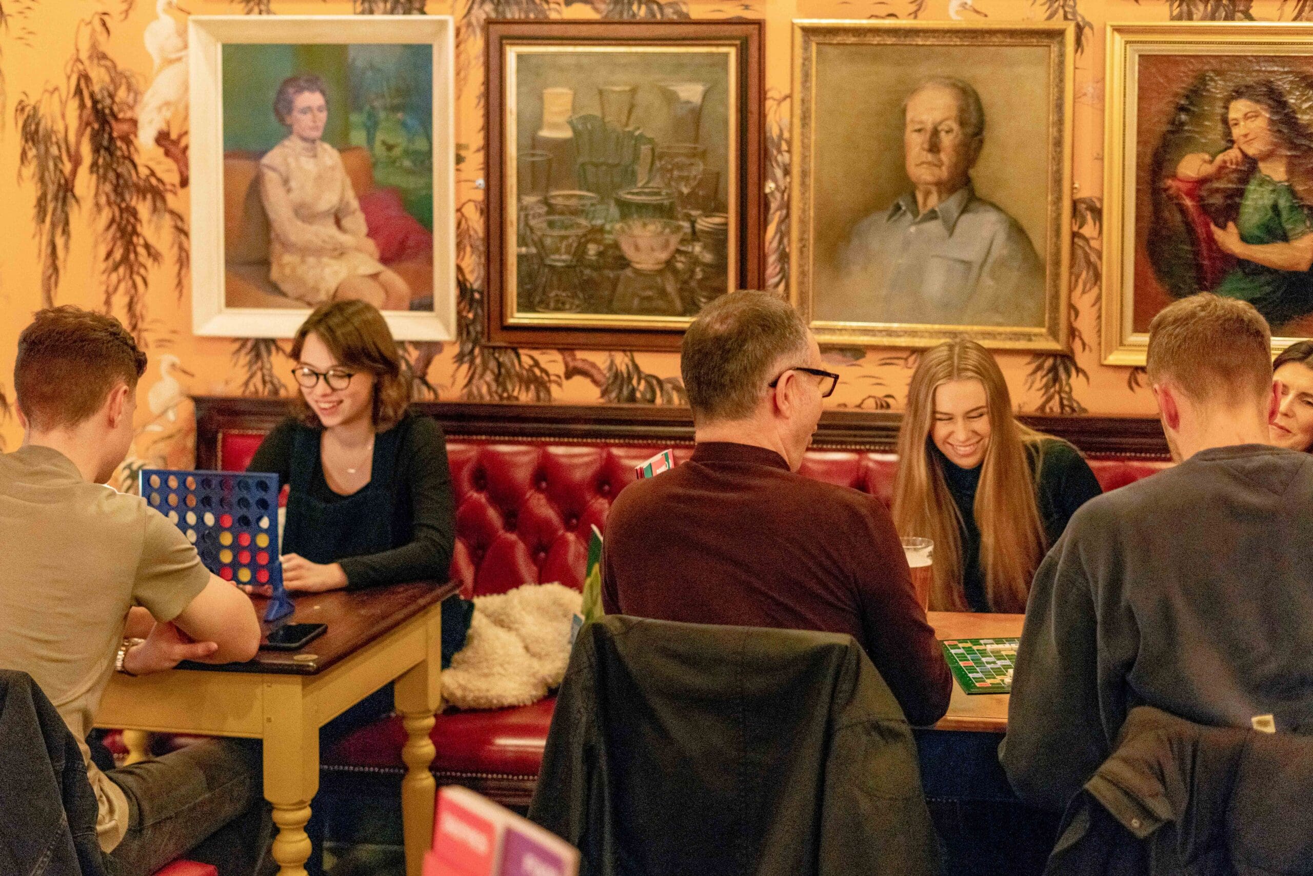 A group of young adults sit at tables in Portello, a cozy, art-filled cafe, playing board games and laughing together, surrounded by framed paintings on the walls and red cushioned seating.