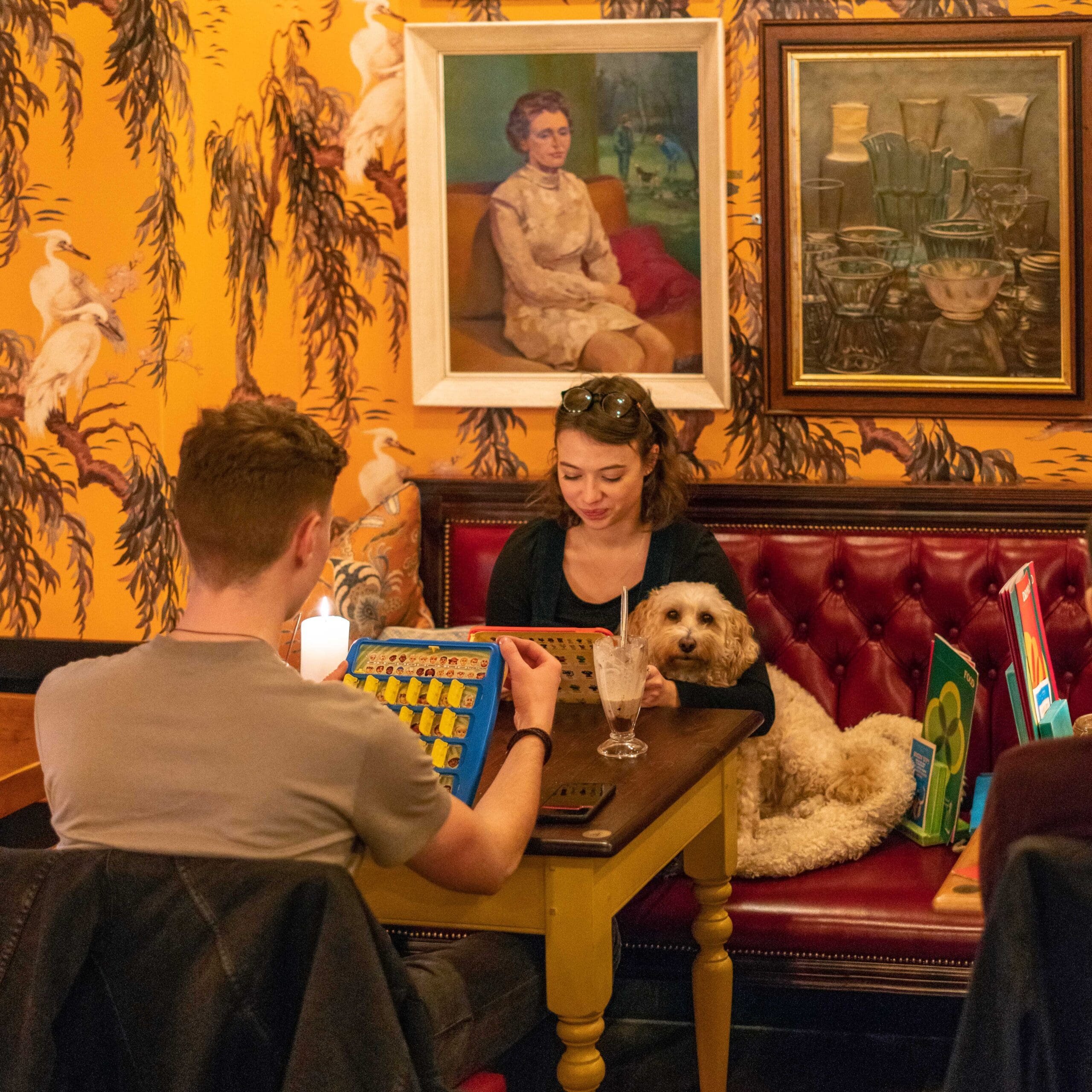 A man and woman play a board game at a café table at Portello while a fluffy dog sits beside them on a red bench. Colorful art and patterned wallpaper decorate the cozy, warmly lit space.