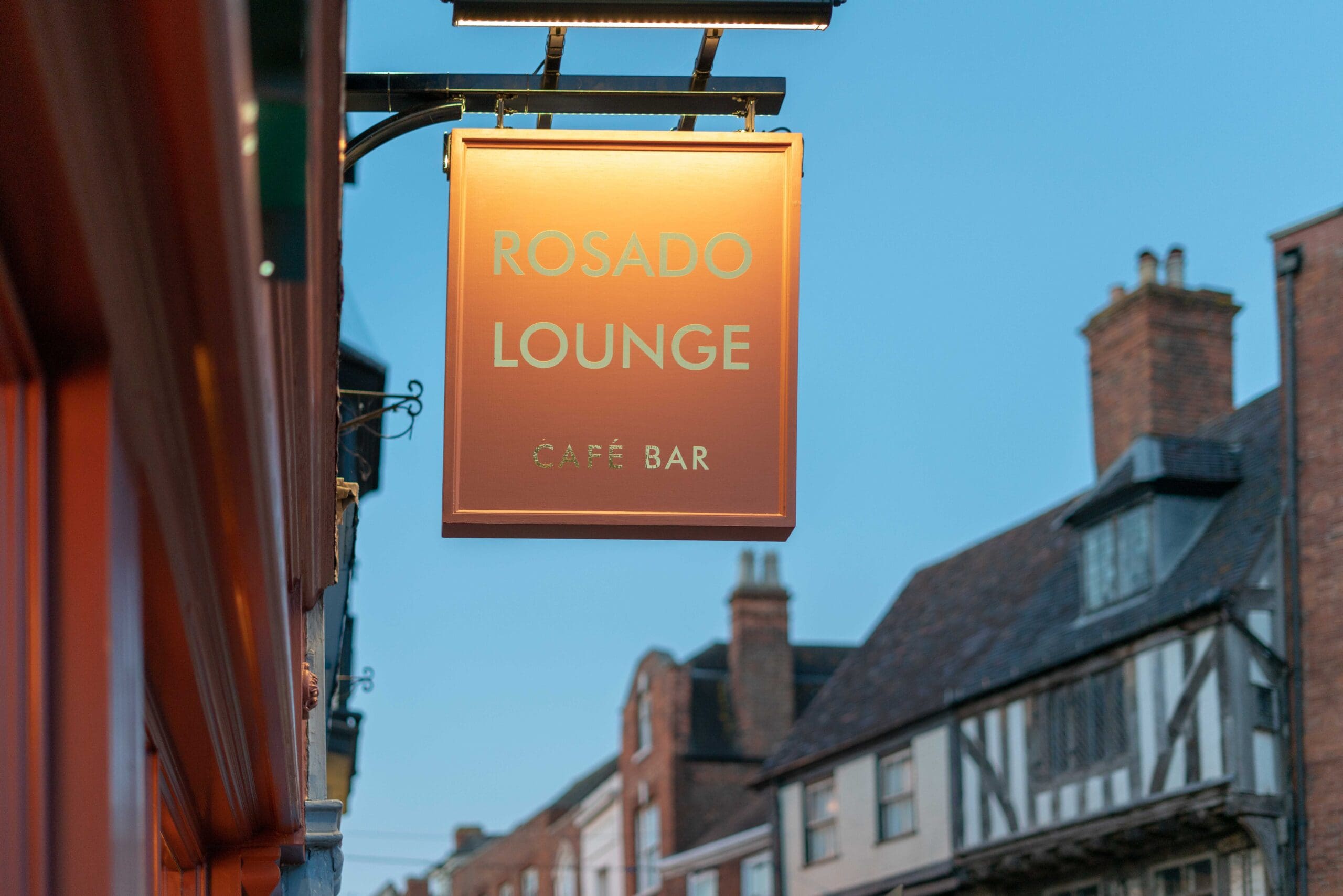 A glowing orange Rosado Lounge Café Bar sign hangs outside a building on a street lined with historic houses under a clear blue sky.