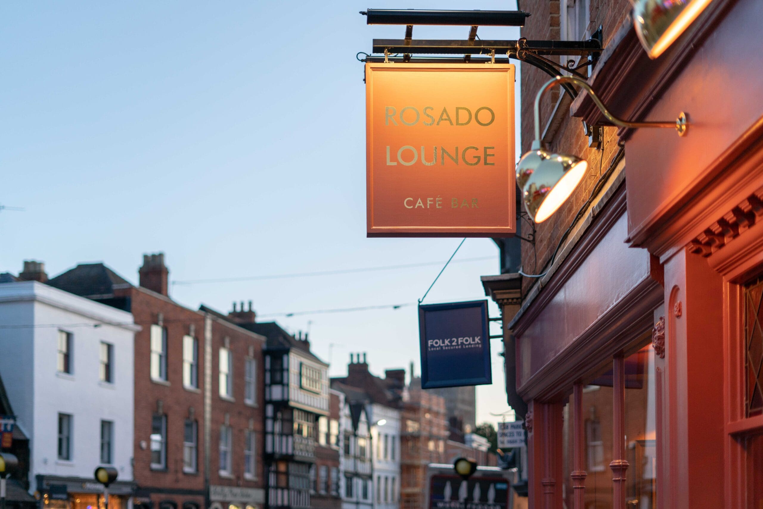 Street view of a row of buildings with a lit Rosado Lounge Cafe Bar sign in the foreground and another sign for "Folk2Folk" further down the street, all beneath an early evening sky.