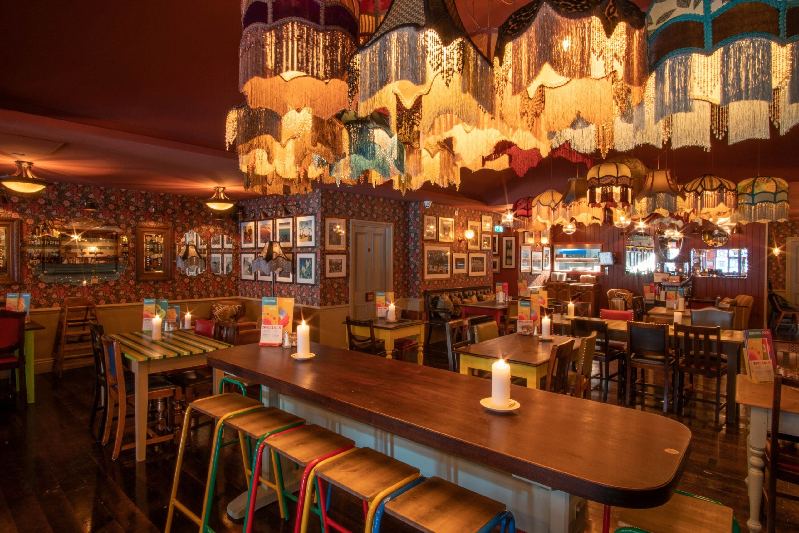 Warmly lit restaurant interior with eclectic, fringed lampshades and colorful Rosado chairs, wooden tables, candles, patterned wallpaper, and framed pictures on the walls, creating a cozy, vintage atmosphere.