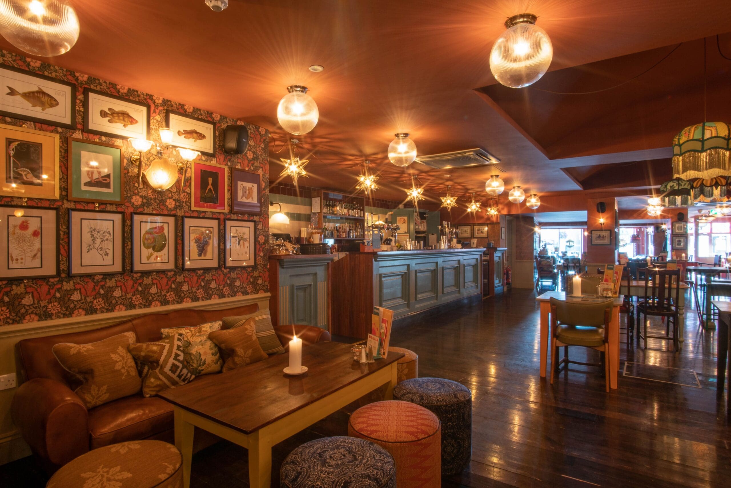 Cozy bar interior with warm lighting, eclectic wall art, comfortable seating including a brown leather sofa and colorful stools, wooden floors, and a long counter in the background. Tables are set for dining—perfect for sipping Rosado with friends.