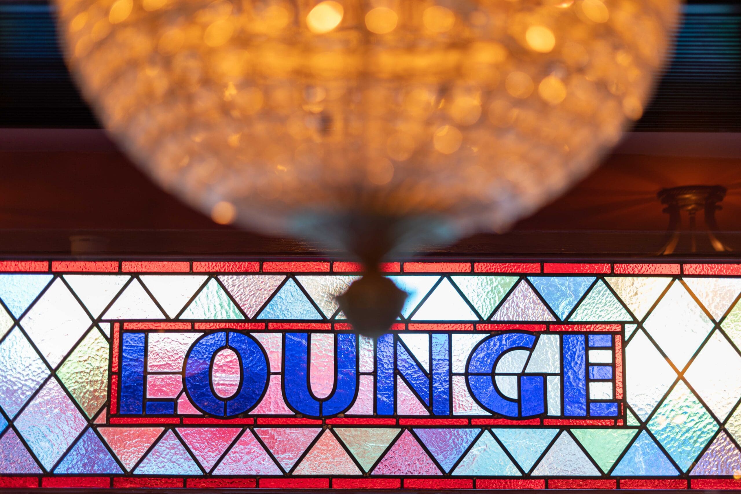 A stained glass window with the word "LOUNGE" in bold blue letters, bordered by a rich rosado hue, with a blurry chandelier hanging above in the foreground.