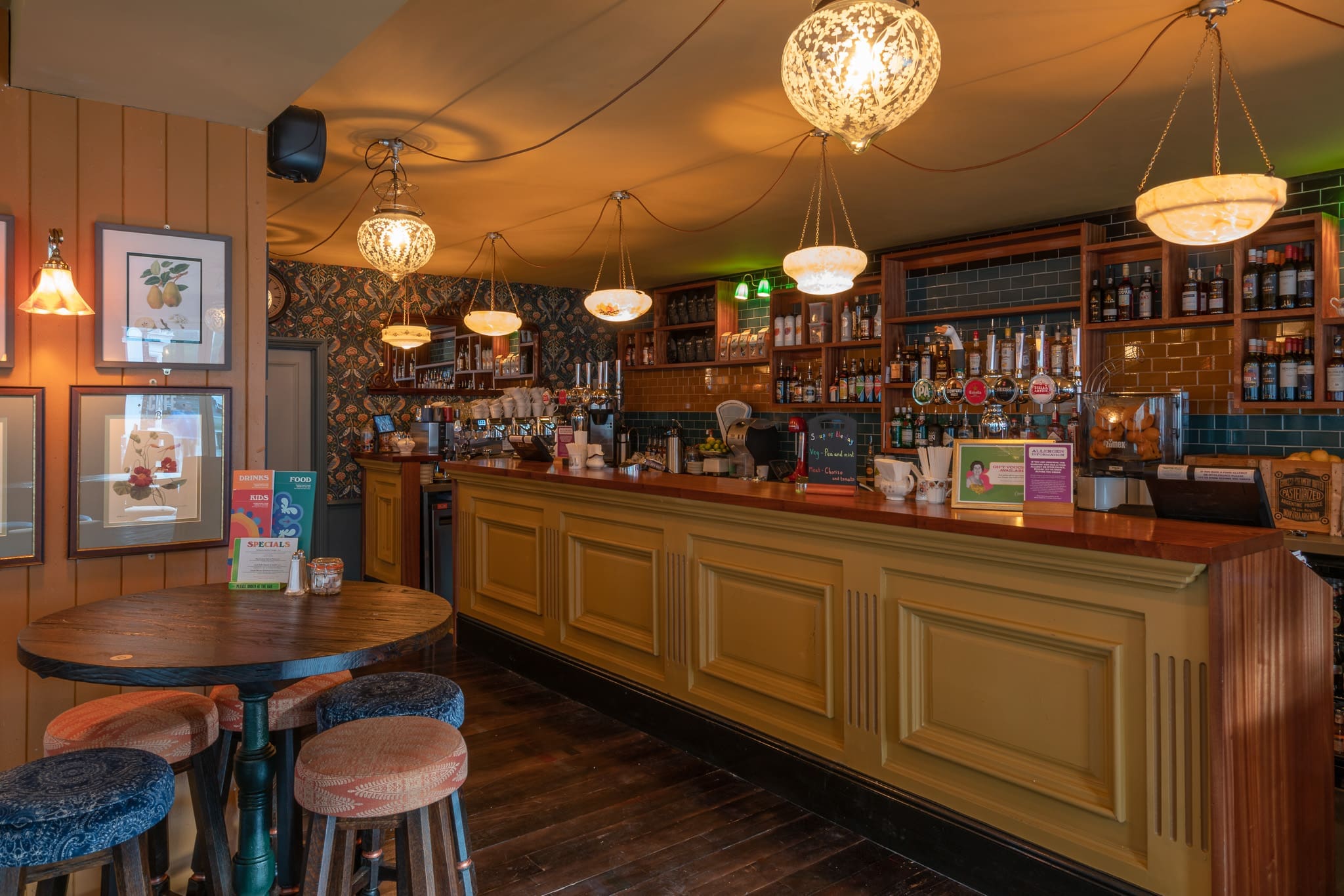 A cozy Sacro pub interior with a long yellow bar, shelves of bottles and glasses, hanging globe lights, and patterned wallpaper. A round wooden table with four upholstered stools sits in the foreground.