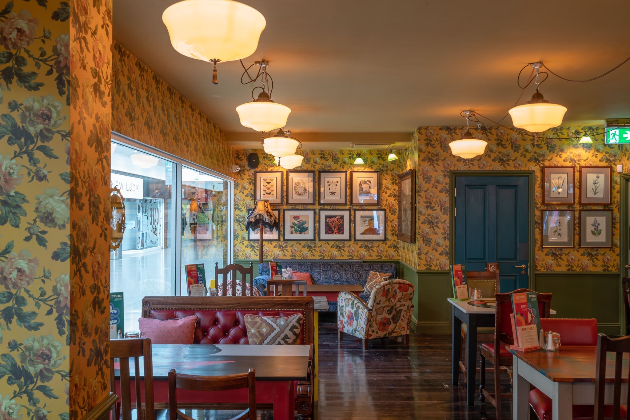 A cozy café interior with floral wallpaper, framed pictures on the walls, vintage-style lamps, wooden tables, cushioned chairs, and a window letting in sacro natural light from the street.