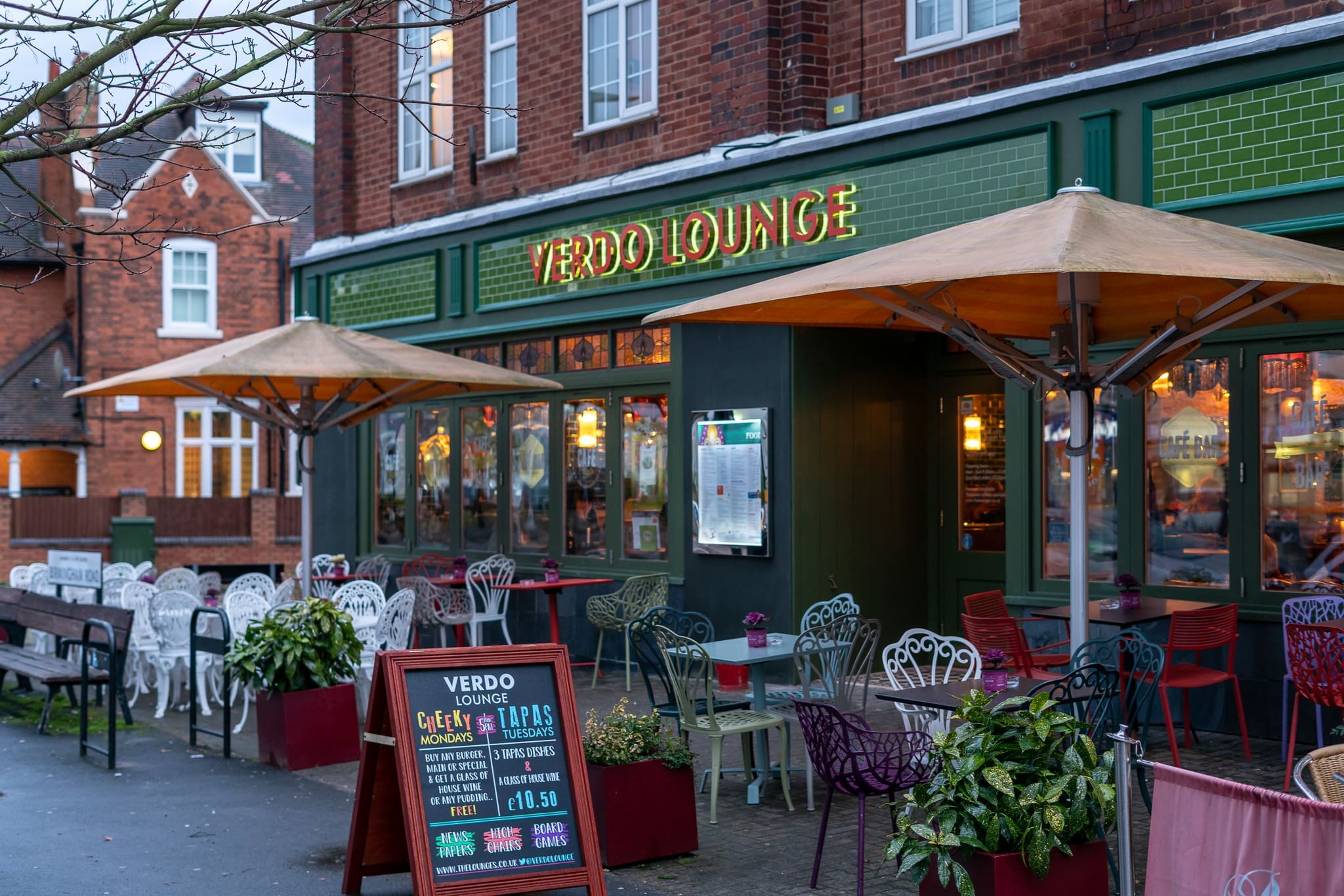 The outdoor seating area of Verdo Lounge café features colorful chairs and tables under umbrellas, vibrant plants in planters, and a Verdo menu board set on the pavement before a charming brick building on a cloudy day.