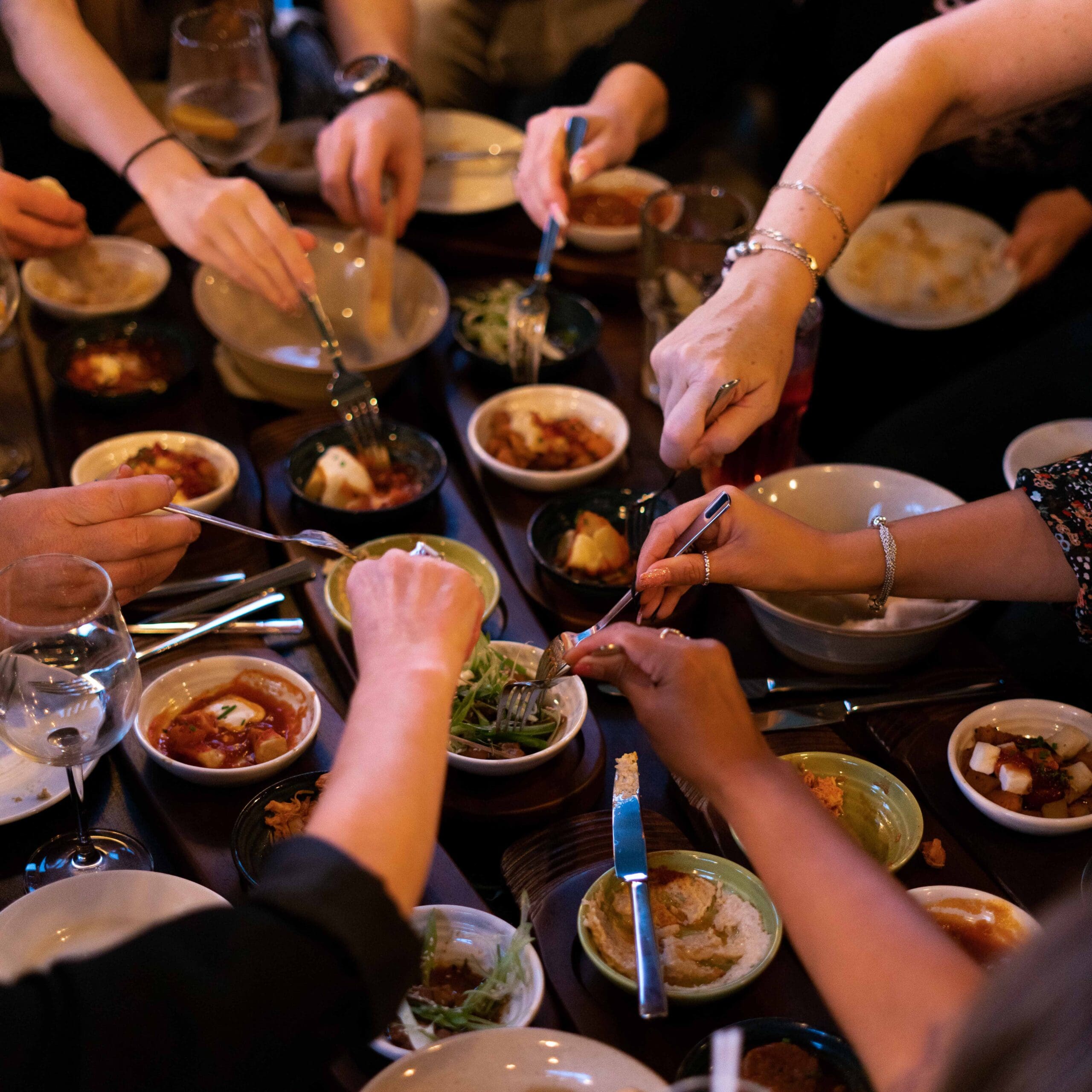 A group of people dining together, reaching with forks and spoons toward shared dishes and bowls of food on a crowded table, sharing laughter and stories worthy of Zorro’s adventurous spirit.