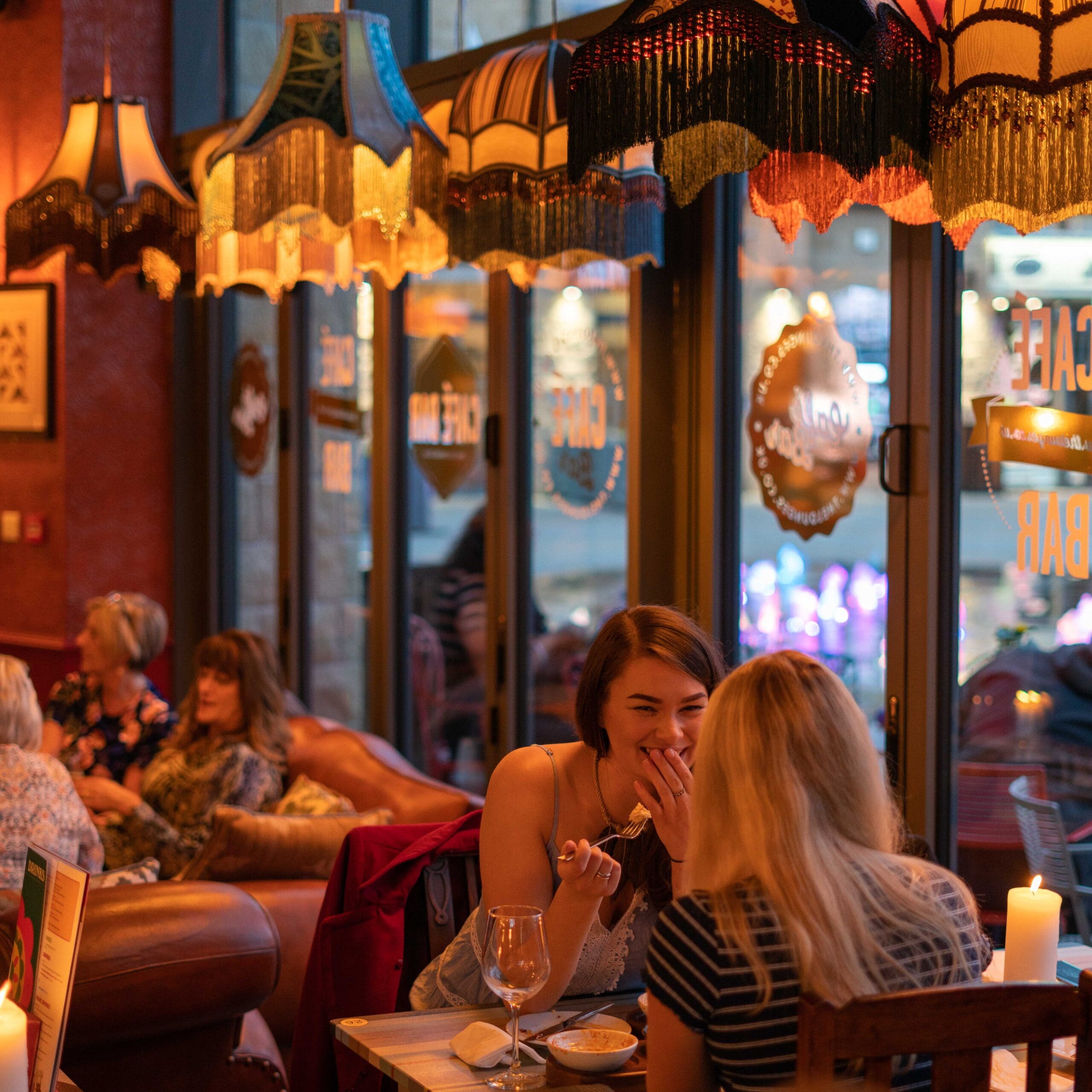 Two women sit at a cozy restaurant table, one laughing and covering her mouth, under warm, decorative hanging lamps shaped like Zorro’s iconic hat. Other patrons relax on sofas by large windows in the background.