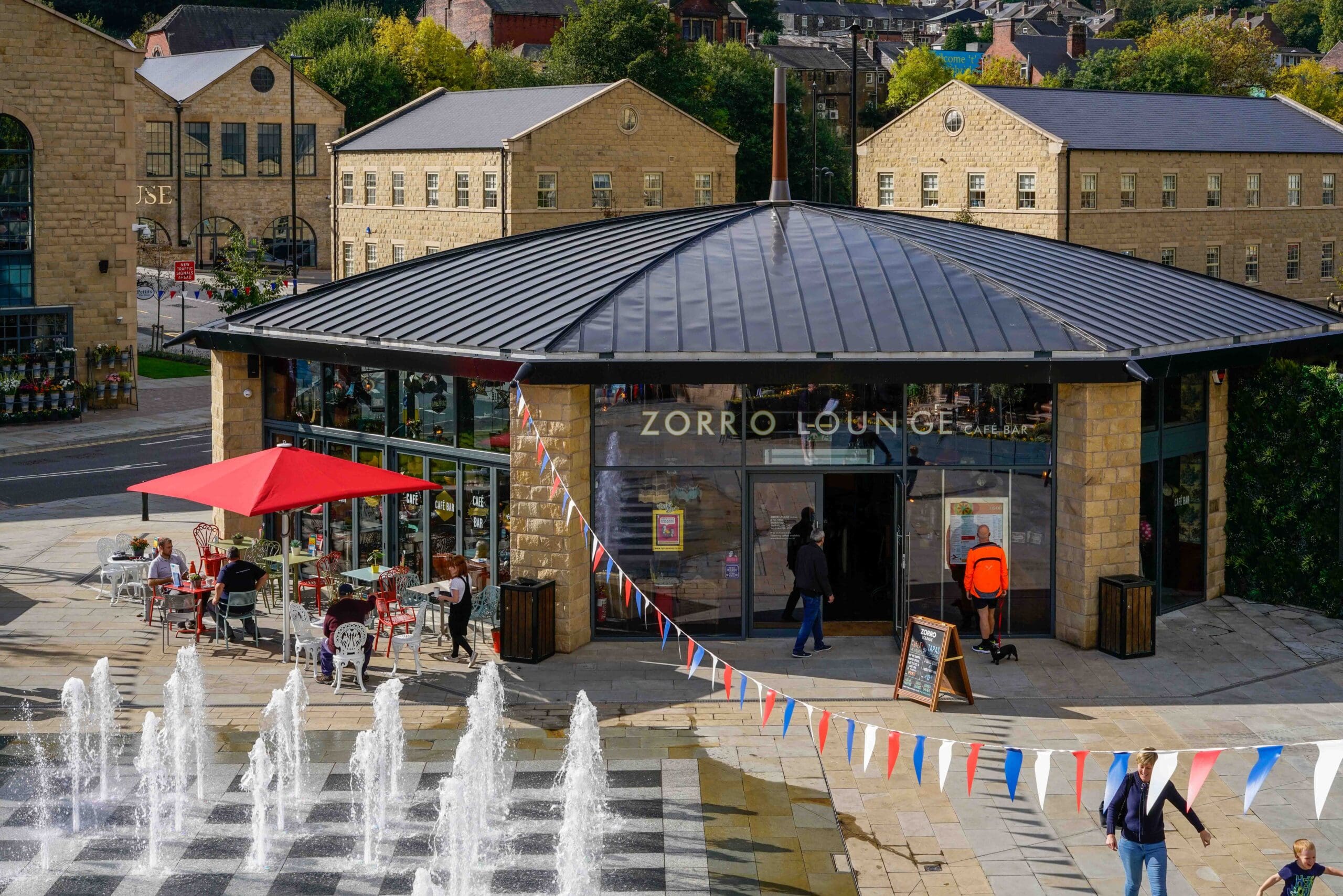 People walk near a circular cafe with glass walls and a sign reading "Zorro Lounge." Red umbrellas shade outdoor tables, and colorful bunting hangs above. Water jets from a Zinco fountain spray in the foreground. Stone buildings are in the background.