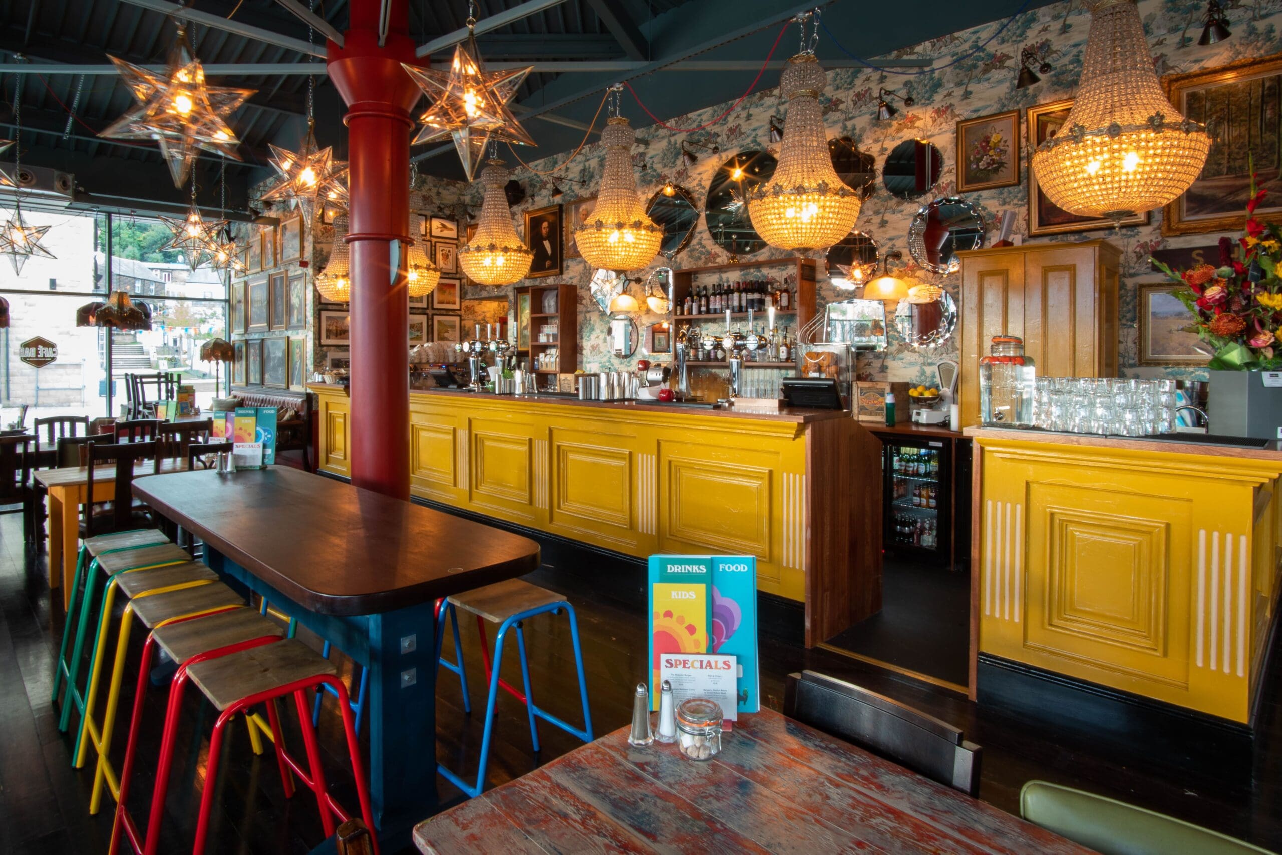 A vibrant bar with a yellow counter, colorful stools, hanging chandeliers, and star-shaped lights. Eclectic wall decor—mirrors, framed art, and floral wallpaper—creates a cozy Zorro-inspired vibe in this lively space with tables and menus visible.