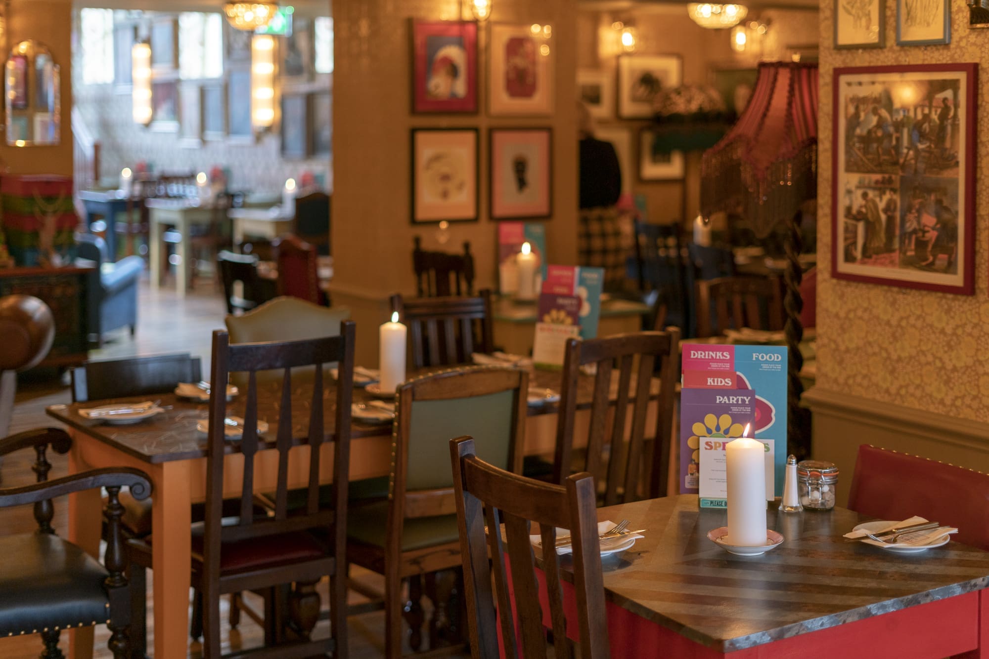 A cozy restaurant interior with wooden tables and chairs, lit candles on each table, colorful menus, framed art on the walls, and warm ambient lighting creates a welcoming fondo perfect for an inviting and relaxed atmosphere.