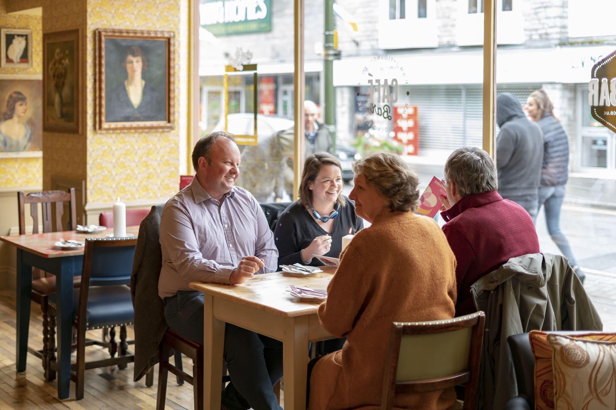 Four adults sit at a table in a cozy café, smiling and conversing. Large windows reveal a lively fondo of pedestrians walking by outside. The café features warm lighting, eclectic chairs, and framed portraits on the walls.