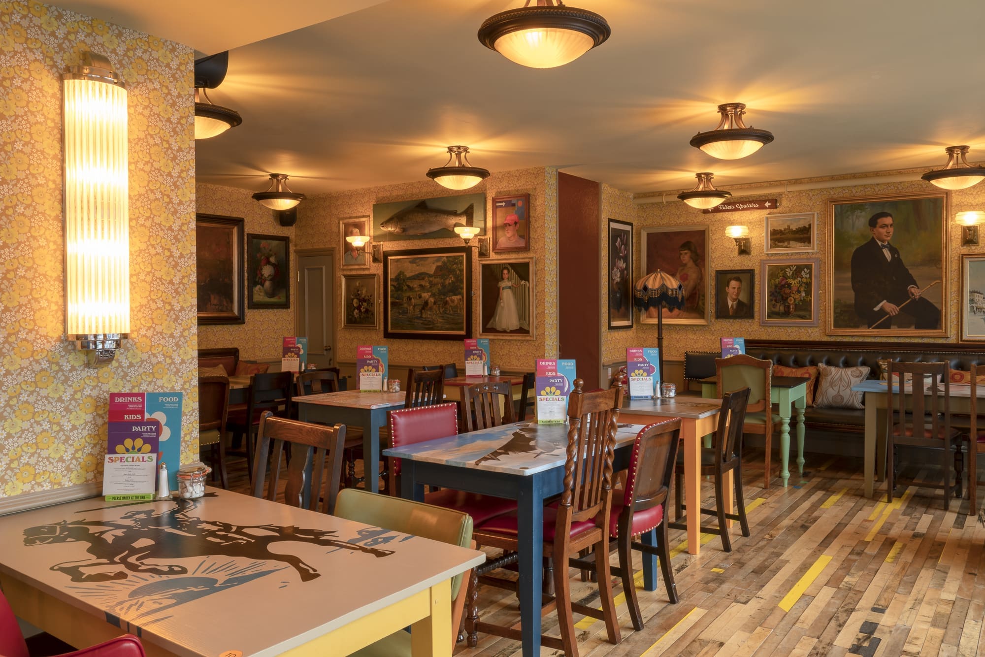 A cozy restaurant interior with wooden tables and chairs, colorful menus on each table, warm lighting, and vintage-style paintings covering the walls creates a Fondo of welcoming, eclectic atmosphere.