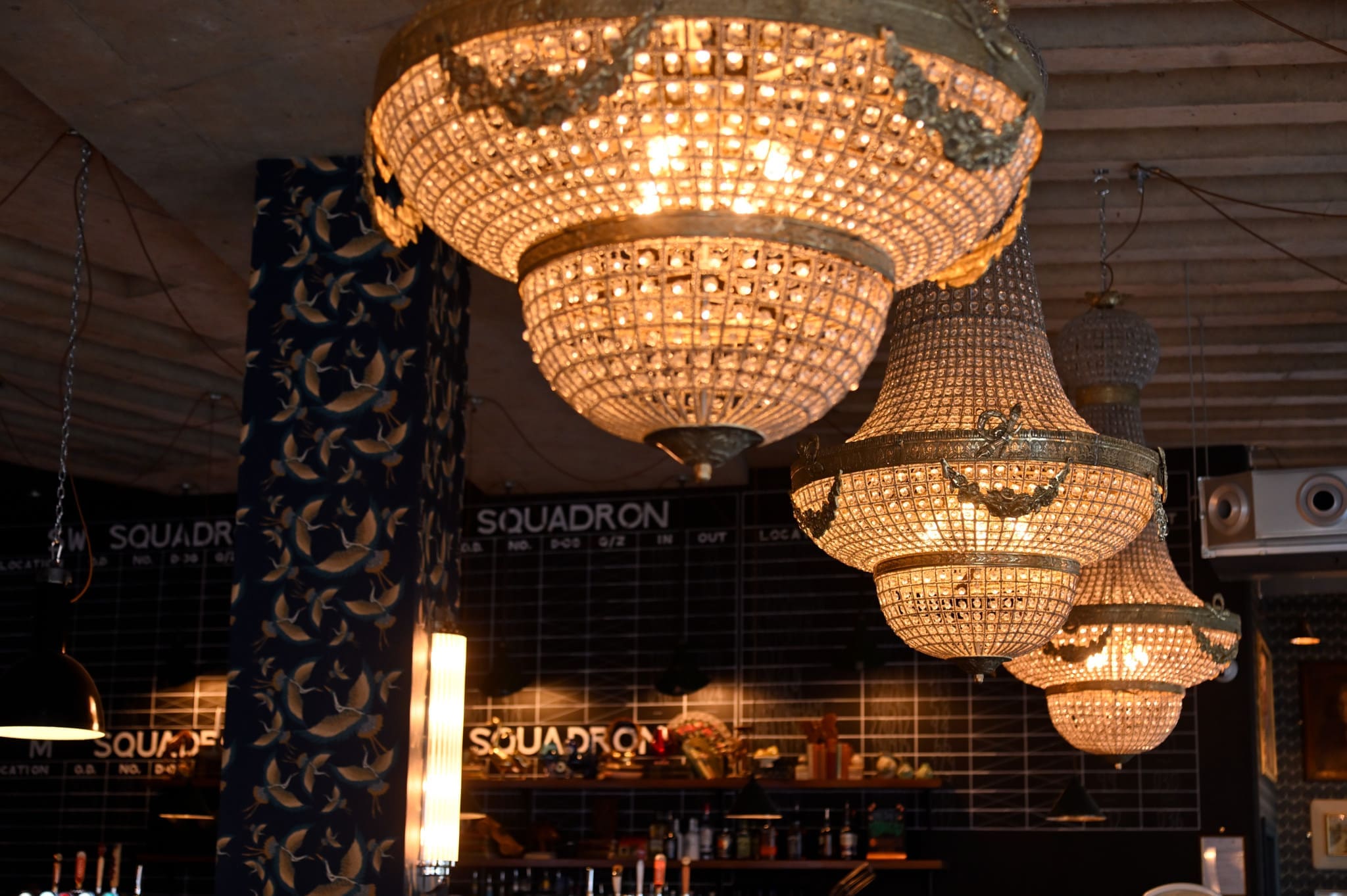 Three ornate crystal chandeliers hang from the ceiling in a stylish, Morello-inspired modern interior with dark tile walls, shelves, and the word "SQUADRON" displayed multiple times in the background.