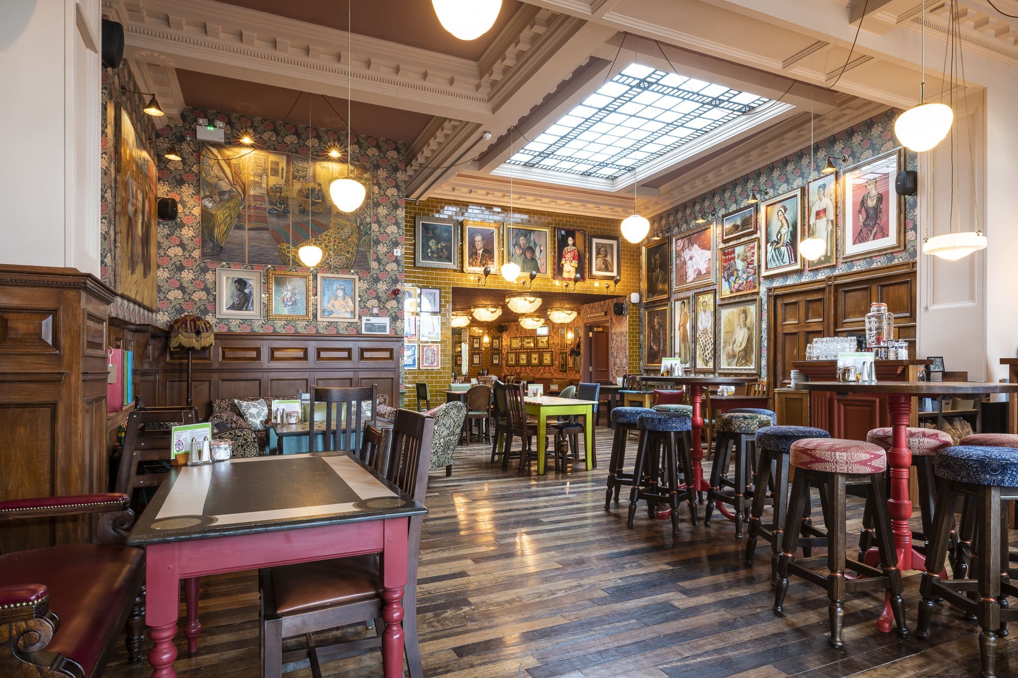 A cozy, vintage-style pub with wooden floors, colorful cushioned stools, and framed artwork covering the walls. Hanging globe lights and floral wallpaper evoke the charm of Morro, while a large skylight brightens dining and socializing spaces.