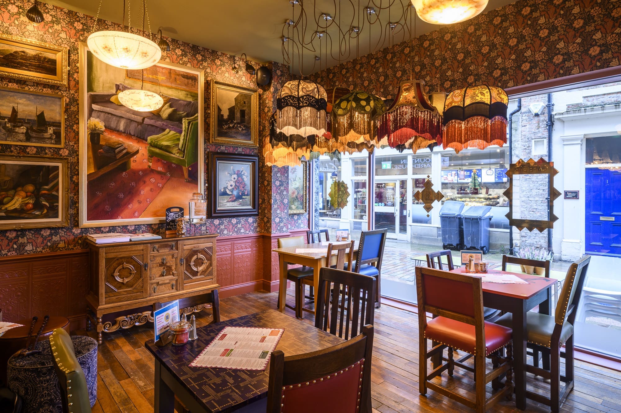 A cozy café with vintage decor, ornate lamps, patterned wallpaper, and framed artwork on the muro. Wooden tables and chairs are arranged near large windows, looking out onto a street with shops.
