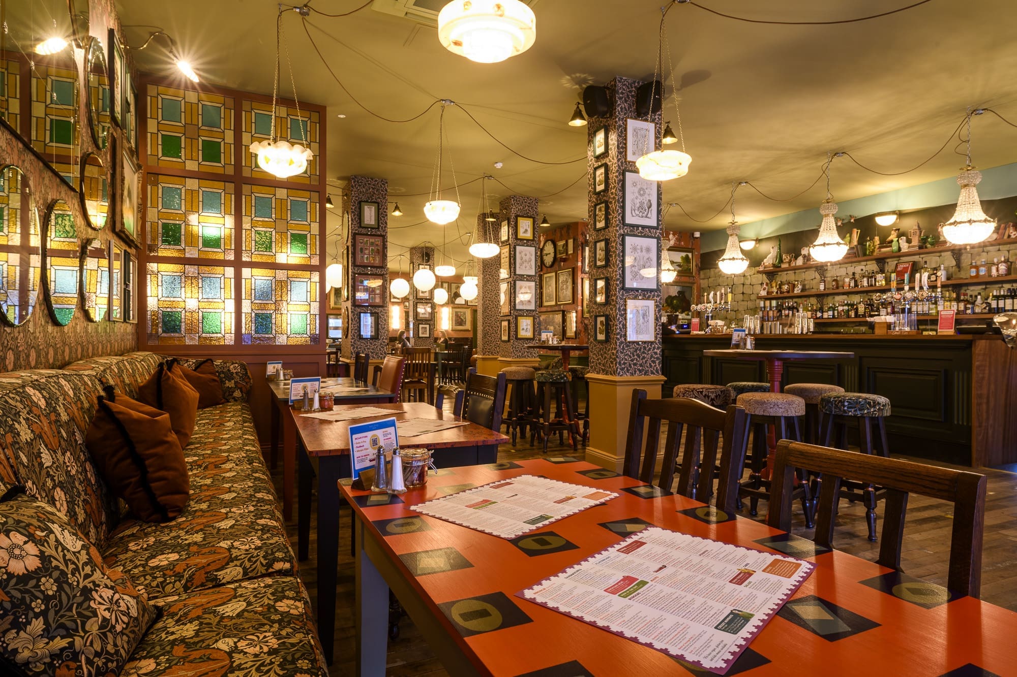Cozy pub interior with patterned sofas, wooden tables set with menus, vintage lighting, stained glass panels, framed pictures on the muro, and a well-stocked bar with high stools in the background.
