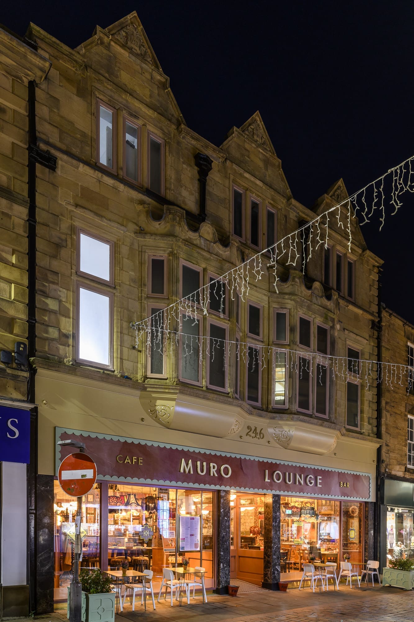 A warmly lit café and bar called "Muro Lounge" on the ground floor of a stone building at night, with string lights above and muro-inspired décor, featuring inviting tables and chairs set outside.
