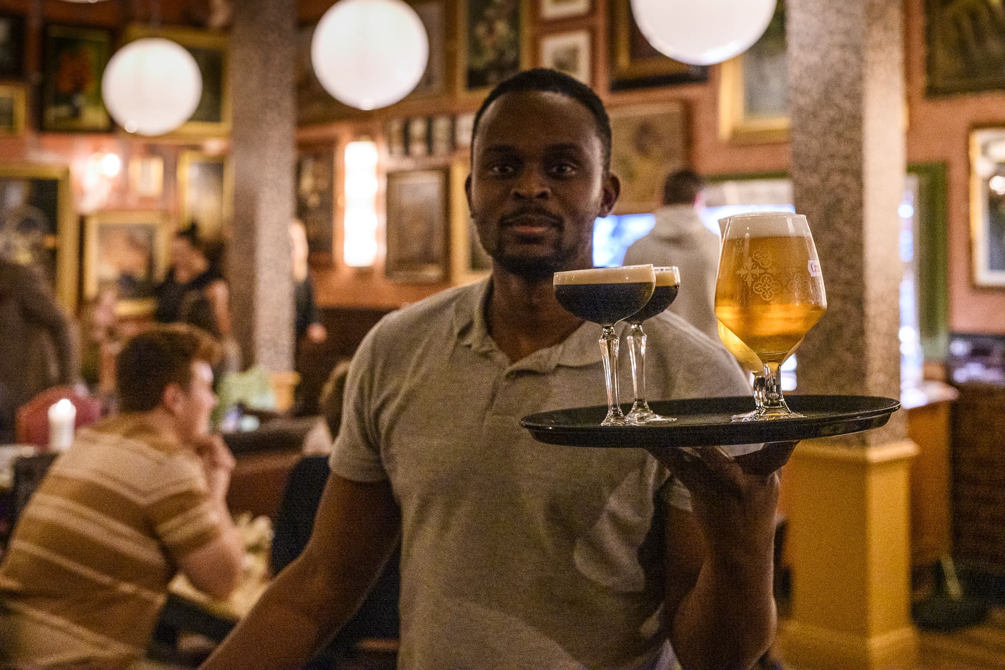 A waiter in a gray polo shirt holds a tray with two drinks—a dark cocktail in a coupe glass and a light beer in a stemmed glass—in a busy, warmly lit bar or restaurant with people and muro décor in the background.