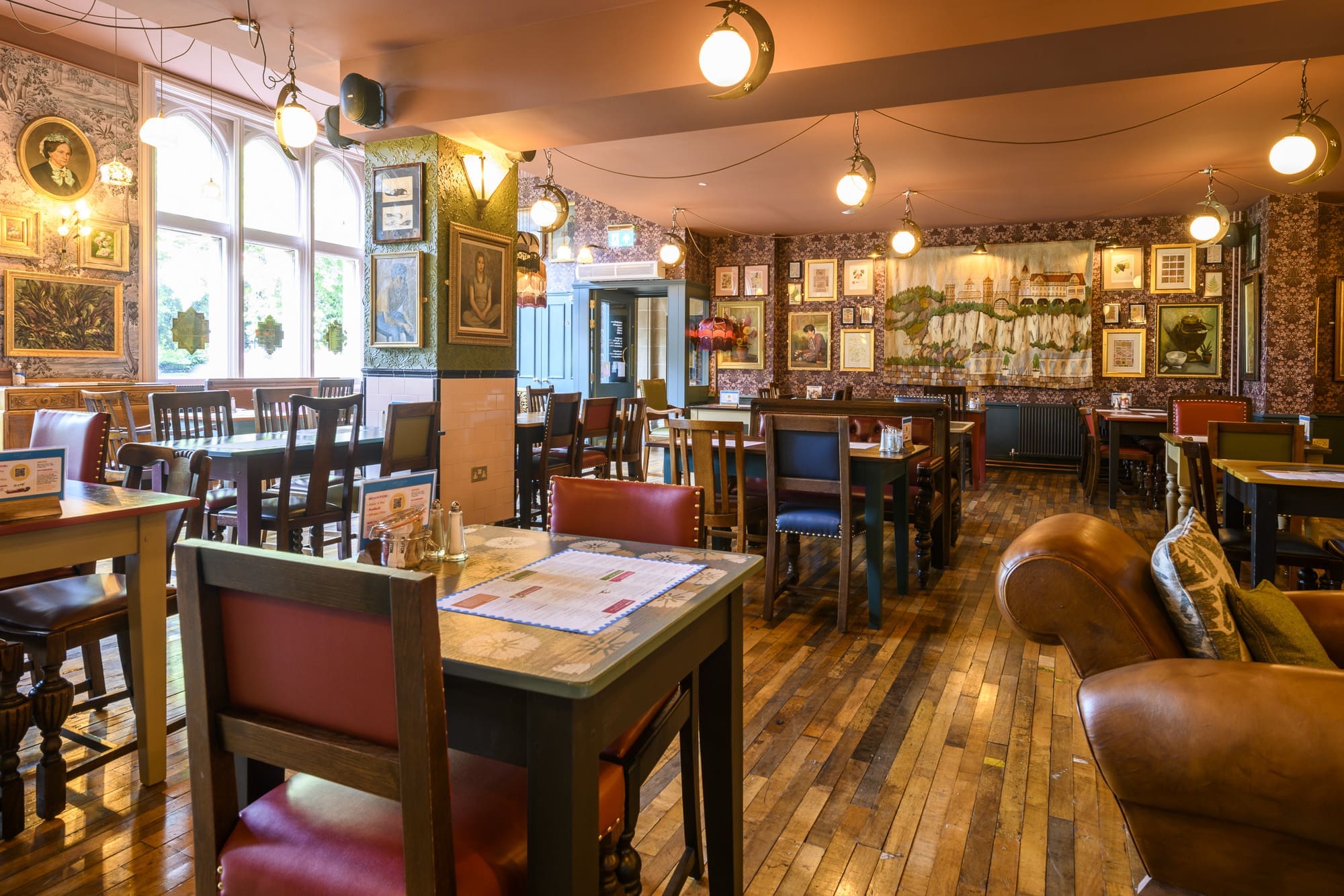 A cozy, eclectic restaurant interior with wooden floors, mismatched chairs, and framed art on patterned Navio walls. Warm hanging lights illuminate tables set for dining as sunlight streams through large arched windows.