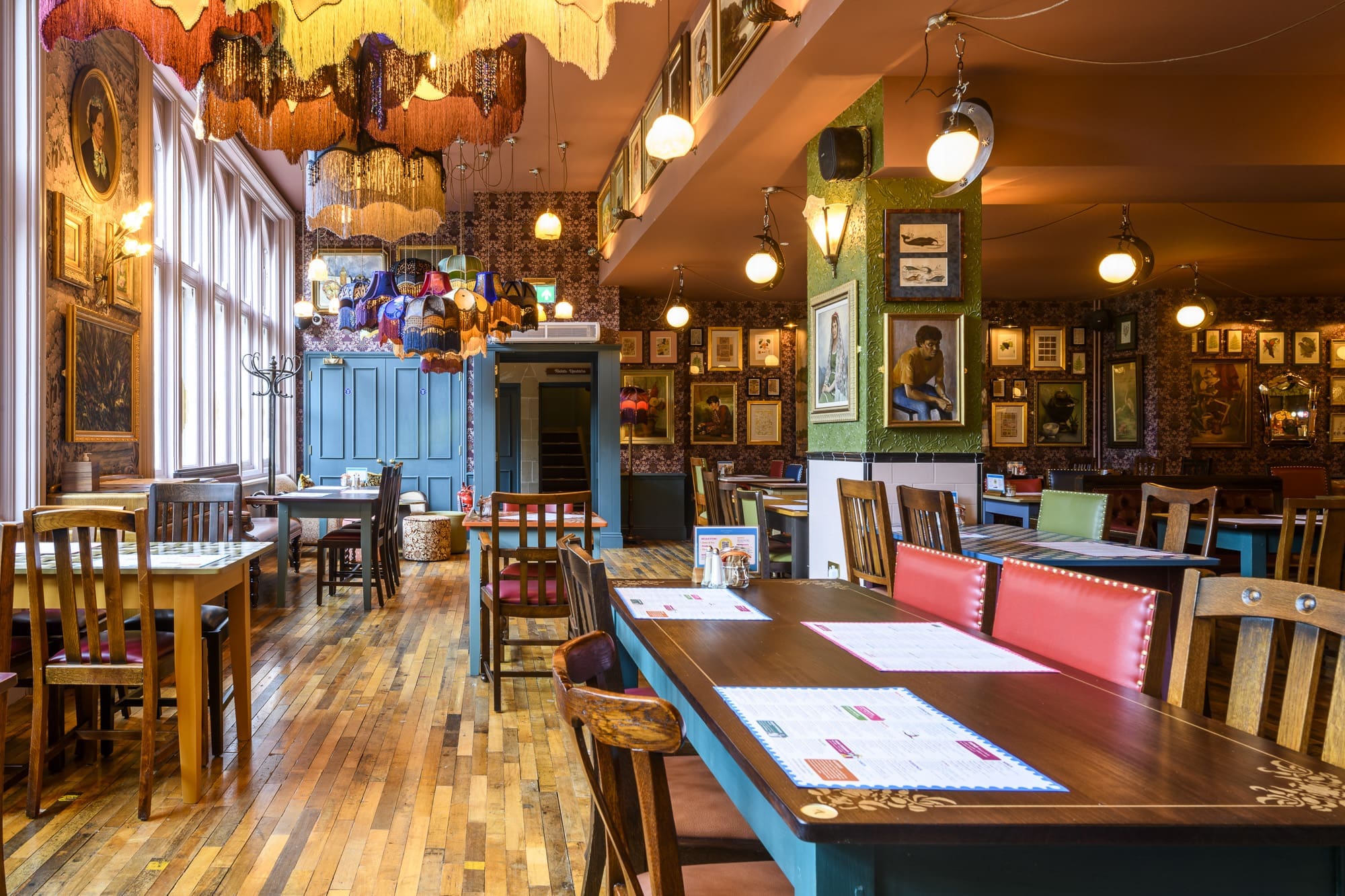 A colorful, eclectic restaurant interior with mismatched wooden chairs, framed art on patterned walls, and Navio pendant lamps. Natural light streams through tall windows onto wooden floors where tables are set with menus and condiments.