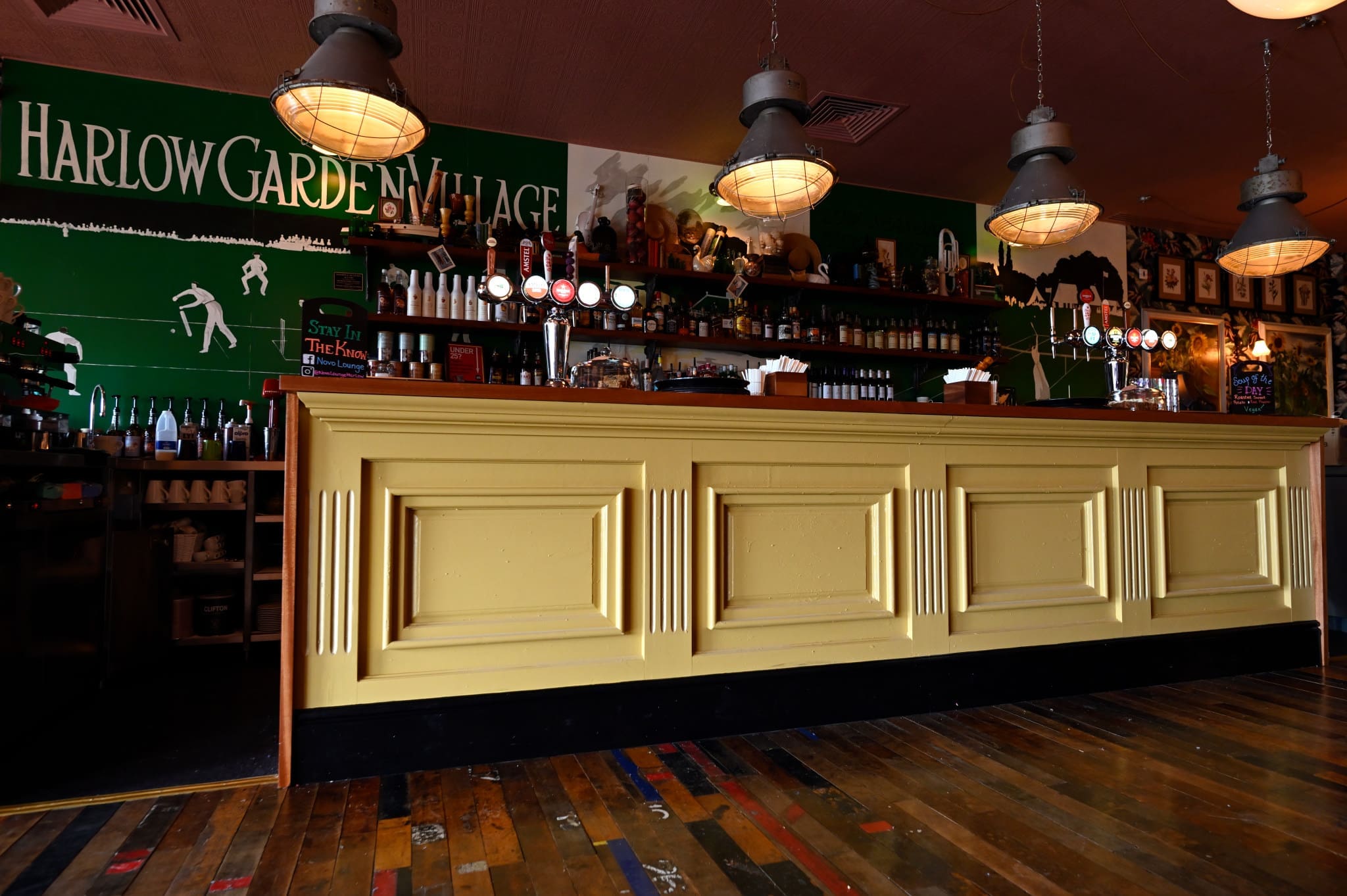 A vintage-style Novo bar features a green wall reading "Harlow Garden Village," a yellow panelled counter, hanging industrial lights, and a variety of bottles and taps on display, all set atop a rustic wooden floor.