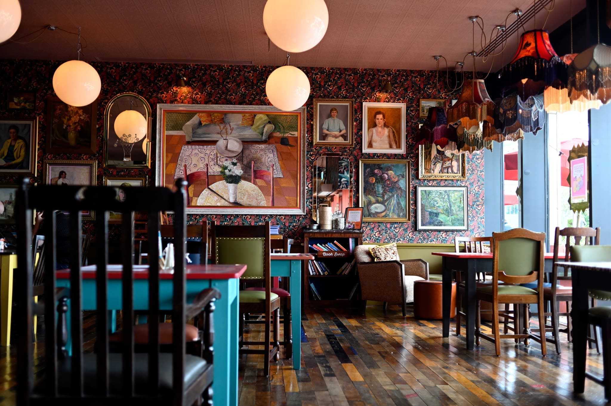A cozy, eclectic café with colorful tables and chairs, wooden floors, and hanging lamps—Novo’s walls are adorned with various framed paintings and portraits, creating a warm, artistic atmosphere.