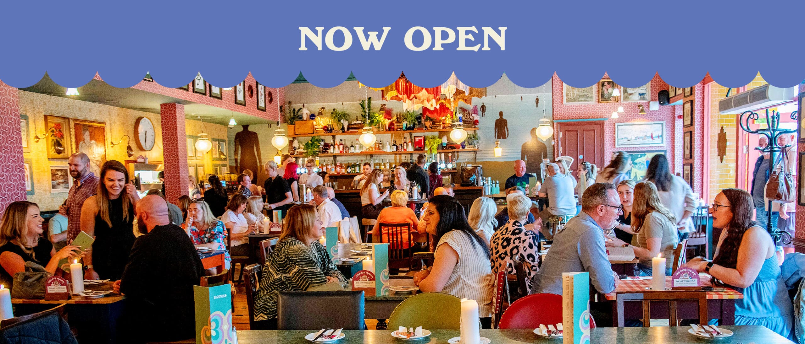 A busy, colorful marinero-themed restaurant filled with people dining and chatting. Staff are serving tables, and the lively atmosphere shines. Text above reads “NOW OPEN” on a blue background with a scalloped edge.
