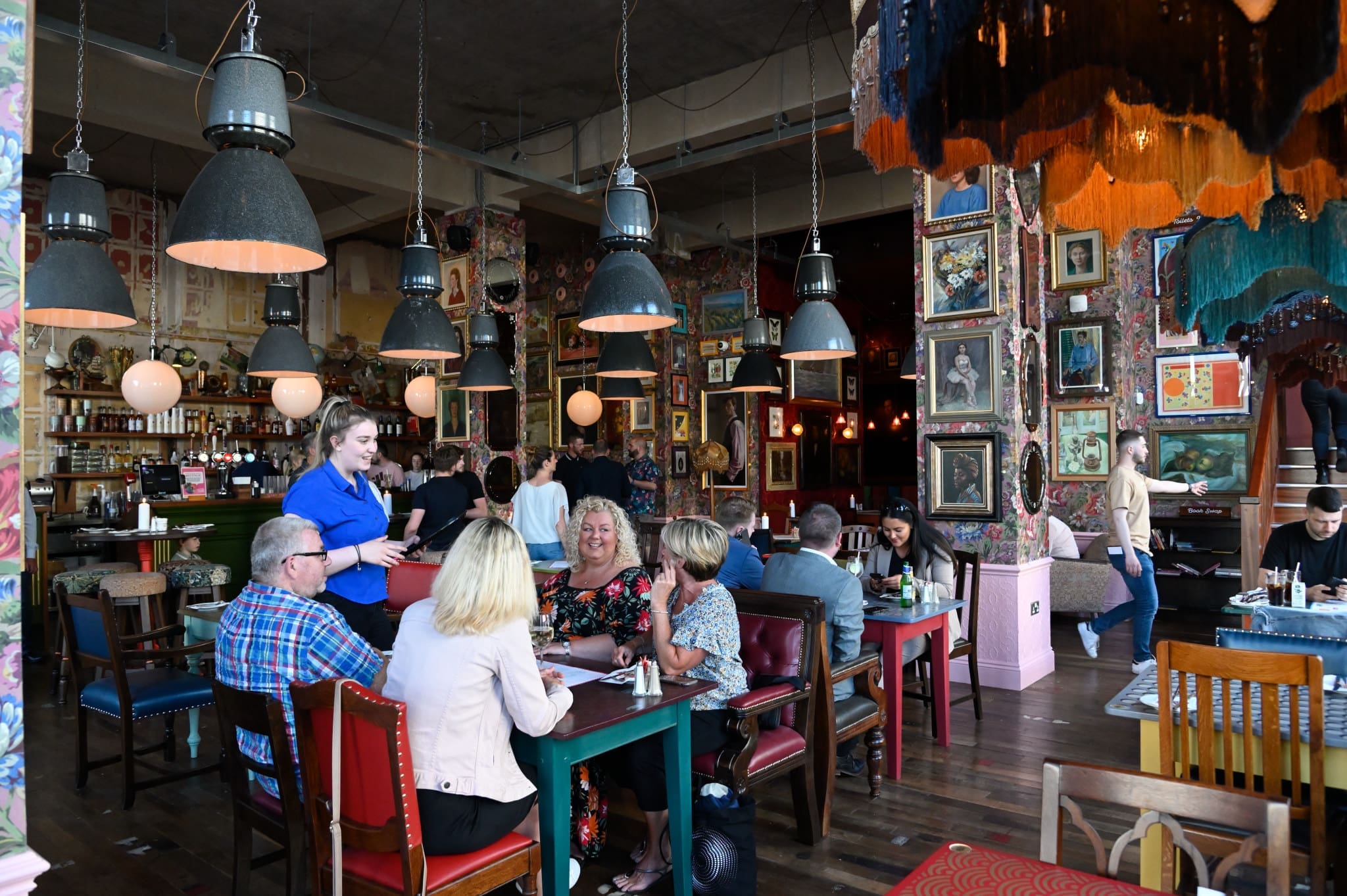 A group of people sit at a colorful table in Occulo, a lively, eclectic restaurant filled with artwork and vintage decor, as a server in blue takes their order. Other diners and staff are visible in the background.