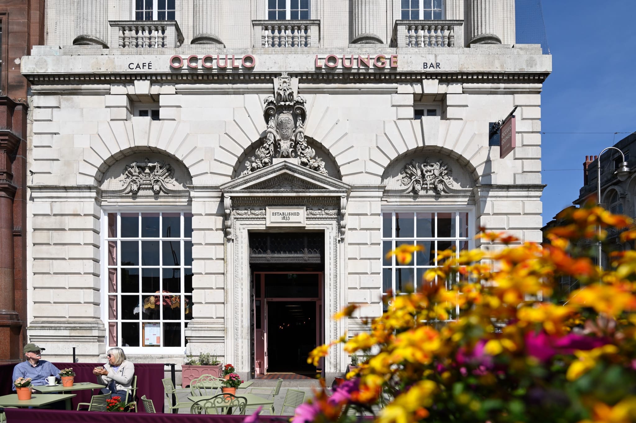 A grand, ornate building with large arched windows houses "Occulo Lounge" café and bar. Two people sit at an outdoor table in front, while colorful flowers blur the foreground under Occulo’s clear blue sky.