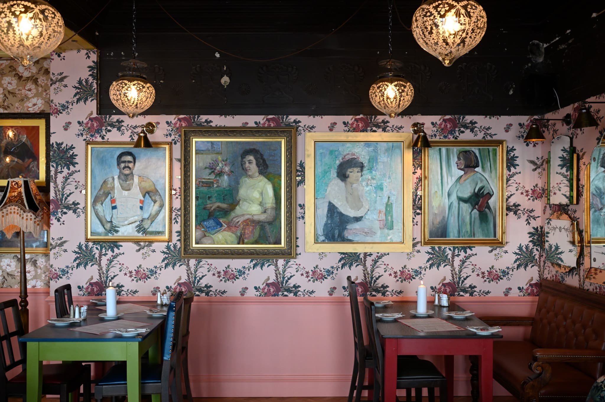 A restaurant with floral wallpaper and four large framed paintings of people on the wall, featuring ornate hanging lights and tables set for dining—a truly Occulo-inspired space.