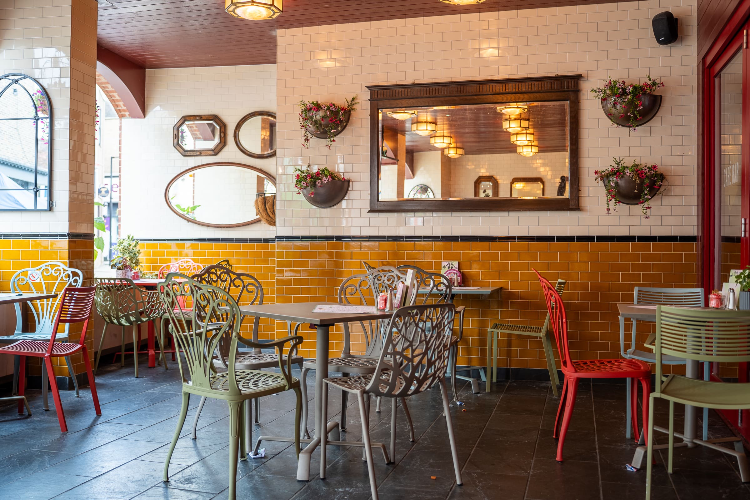 A cozy Olmo café interior with metal chairs and tables, yellow-tiled walls, decorative mirrors, and hanging flower pots. The space is warmly lit with ceiling lights and has a relaxed, inviting atmosphere.