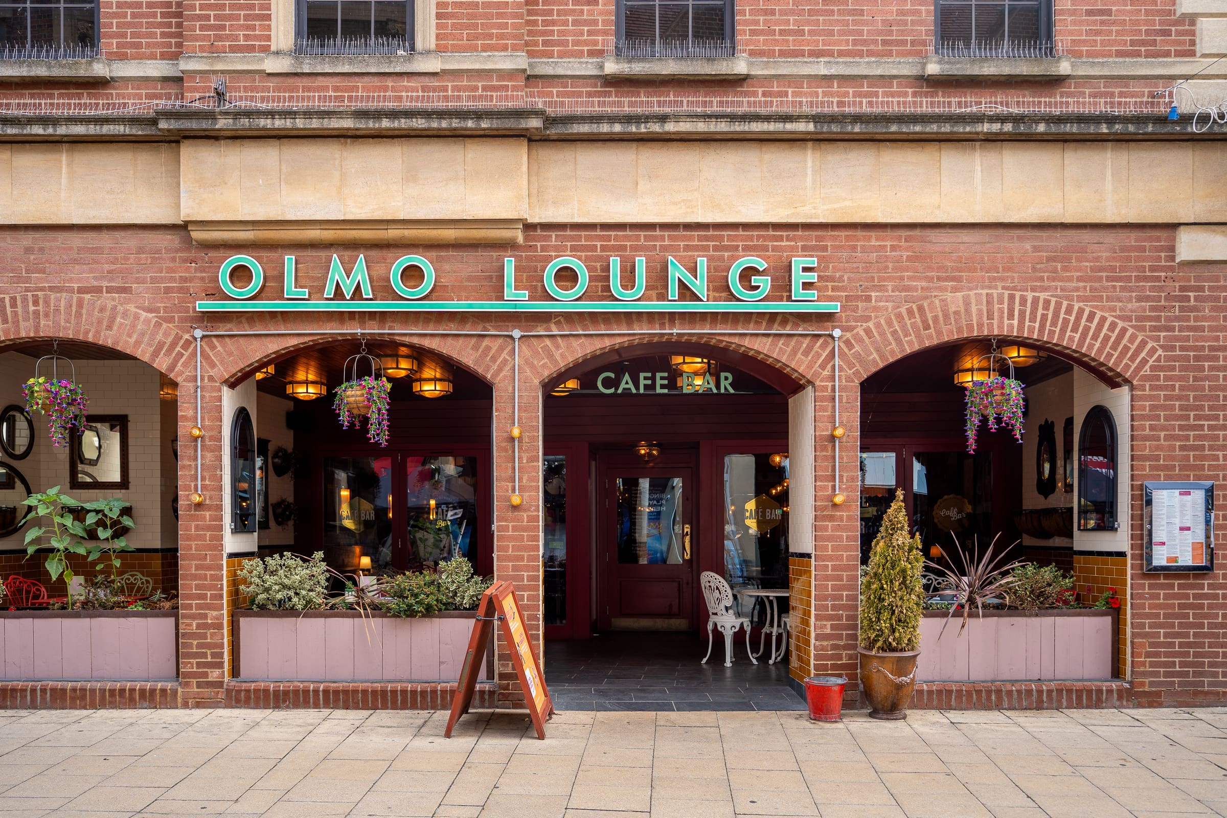 The exterior of Olmo Lounge features large arched windows, lush plants, and a neon Olmo sign above the entrance on a brick building. Outdoor seating and decorative lighting are visible through the windows.