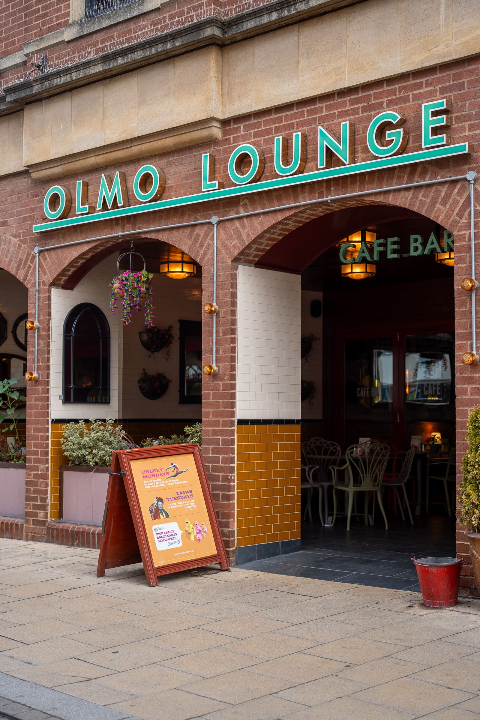 The entrance of Olmo Lounge cafe bar features neon Olmo signage, outdoor seating, lush hanging plants, and a standing signboard displaying promotions on the sidewalk.
