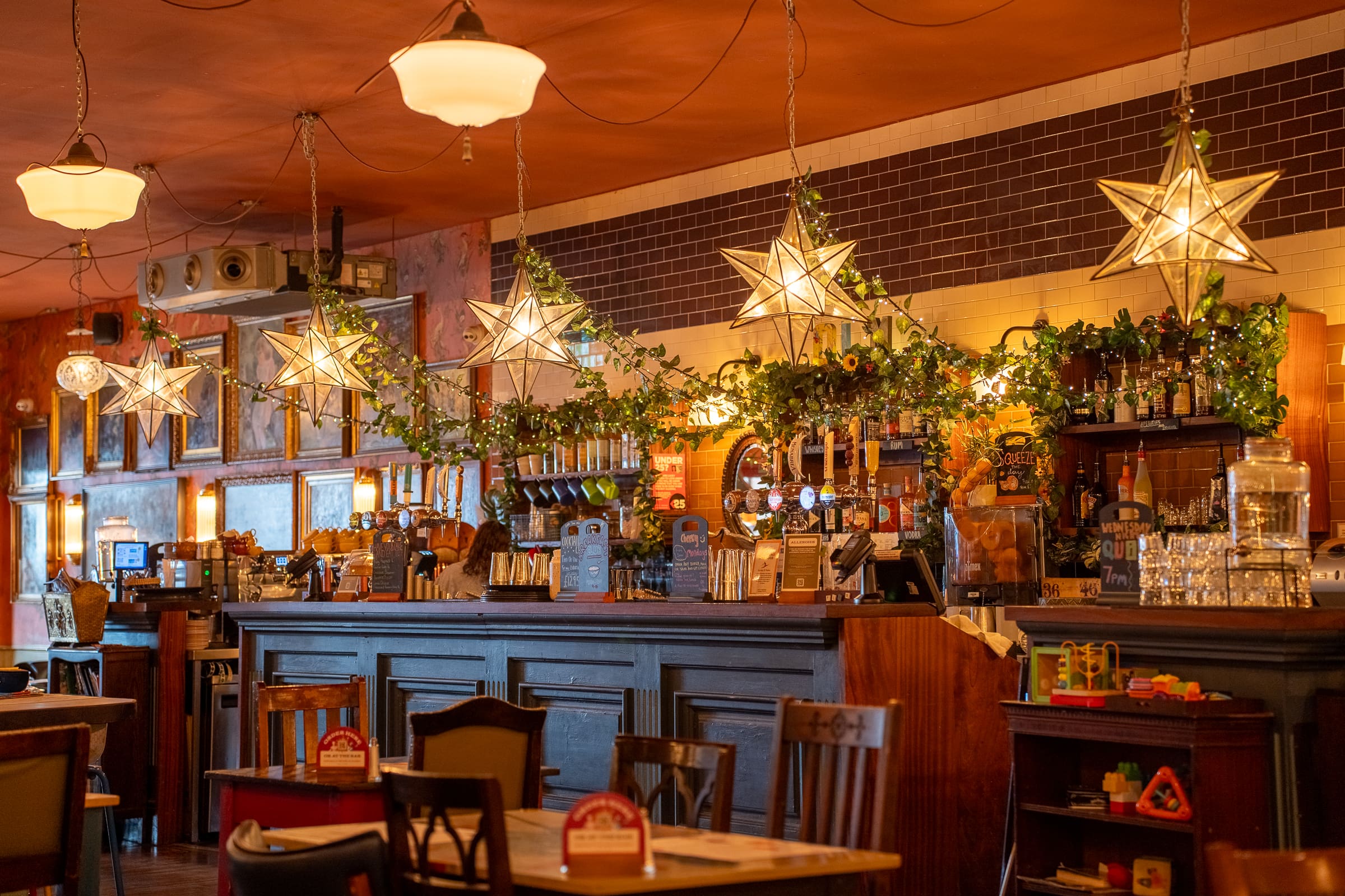 A cozy bar with wooden tables and chairs, illuminated by star-shaped pendant lights. At Olmo, the bar counter is decorated with greenery and bottles, creating a warm and inviting atmosphere.