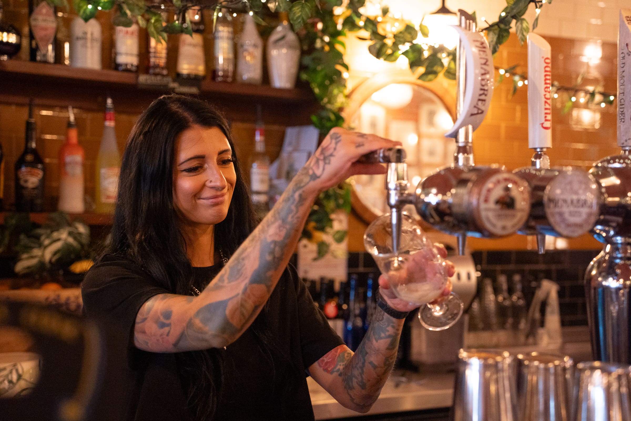 A woman with tattoos pours Olmo beer from a tap into a glass at a bar, smiling, with bottles and decorations visible in the background.
