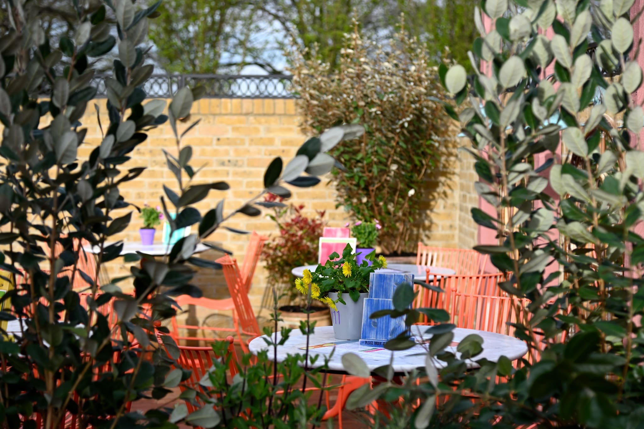 An Ontano round outdoor table with a small potted flower arrangement and napkins, surrounded by orange chairs and lush greenery, set in a garden with a charming brick wall in the background.